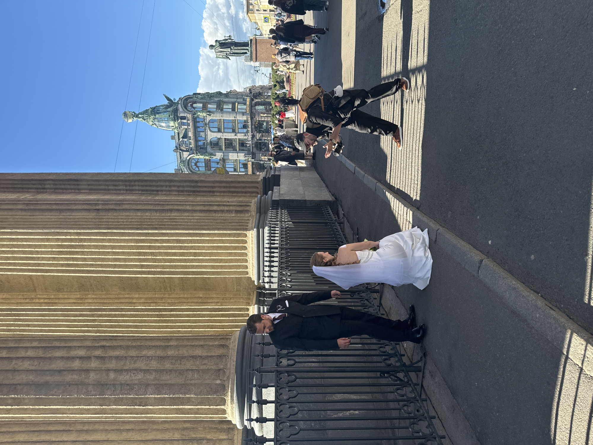 A wedding photoshoot is taking place on a sunny day with a bright blue sky. The subjects are a bride in a white wedding dress and veil and a groom in a black suit and tie. They are posed near a large stone building with columns, likely a historic landmark. In the background, a photographer in a black outfit and a backpack is seen in a dynamic pose, appearing to be capturing the couple's photographs. He is holding a camera and has a white mug attached to his backpack.
Beyond the photographer, a bustling street scene unfolds with pedestrians walking along a sidewalk. Prominent in the background is an ornate building with a distinctive green dome topped by a globe-like structure. The architecture suggests a European city, and the context provided identifies the location as Saint Petersburg, Russia. The time of day appears to be afternoon, judging by the long shadows cast across the street. The weather is clear and sunny, contributing to a festive atmosphere for the wedding celebration. The image captures a candid moment during a wedding photoshoot, blending the romantic occasion with the vibrant urban environment. No visible text is present in the image.