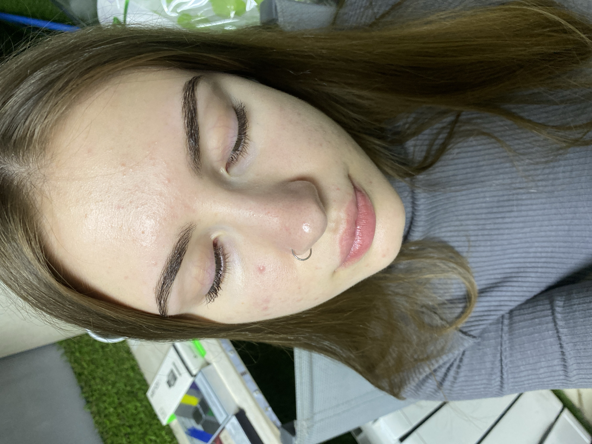 A young woman with brown hair and light skin is photographed from a slightly high angle. She is looking down with her eyes closed, and her head is tilted to the side. She is wearing a grey, ribbed turtleneck sweater. She has a small hoop piercing in her nose. Her face has some visible pores and a few small red blemishes. In the background, there is a blurred white table with some boxes on it, and a green, textured wall. The setting appears to be indoors, possibly a salon or a treatment room. There are no specific indicators of the location being in the city of Дьяково, Russia.