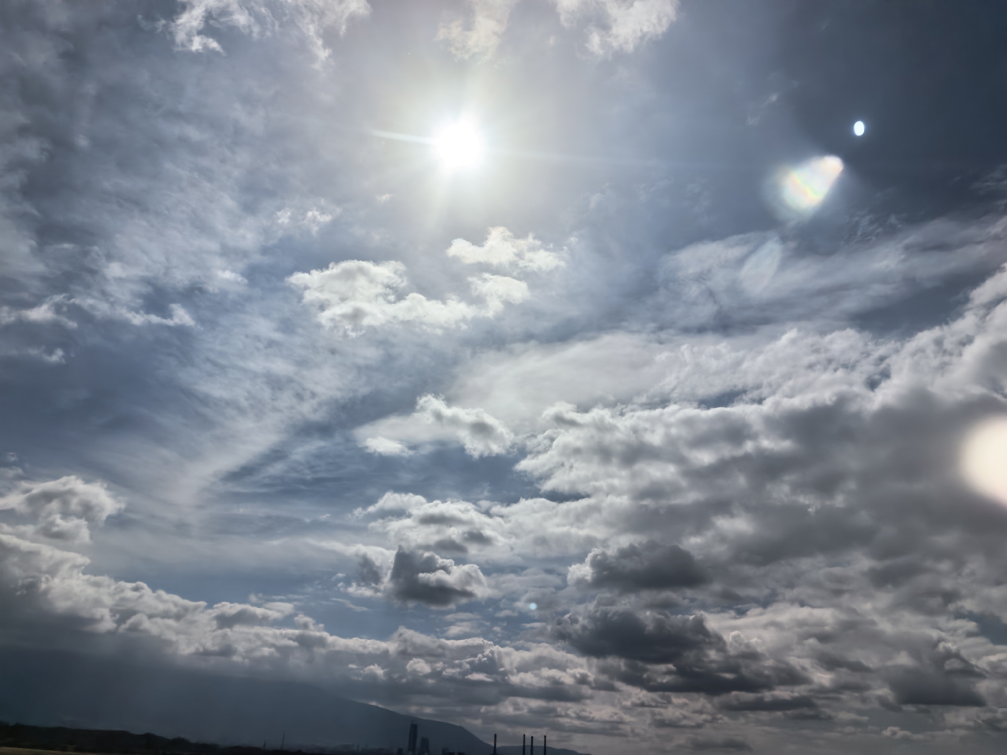 The image captures a dynamic sky over the city of Kazichene, Bulgaria. The sun is bright, peeking through scattered clouds, creating lens flares and highlights. The clouds themselves vary in density, from thin, wispy formations to darker, more substantial masses, suggesting a mixed weather condition. In the distance, a low-lying landscape is visible, with a silhouette of buildings and industrial structures, including what appear to be chimneys, suggesting an urban or industrial area. The scene is devoid of any people or animals. The overall impression is one of a natural landscape juxtaposed with human infrastructure under a dramatic sky.