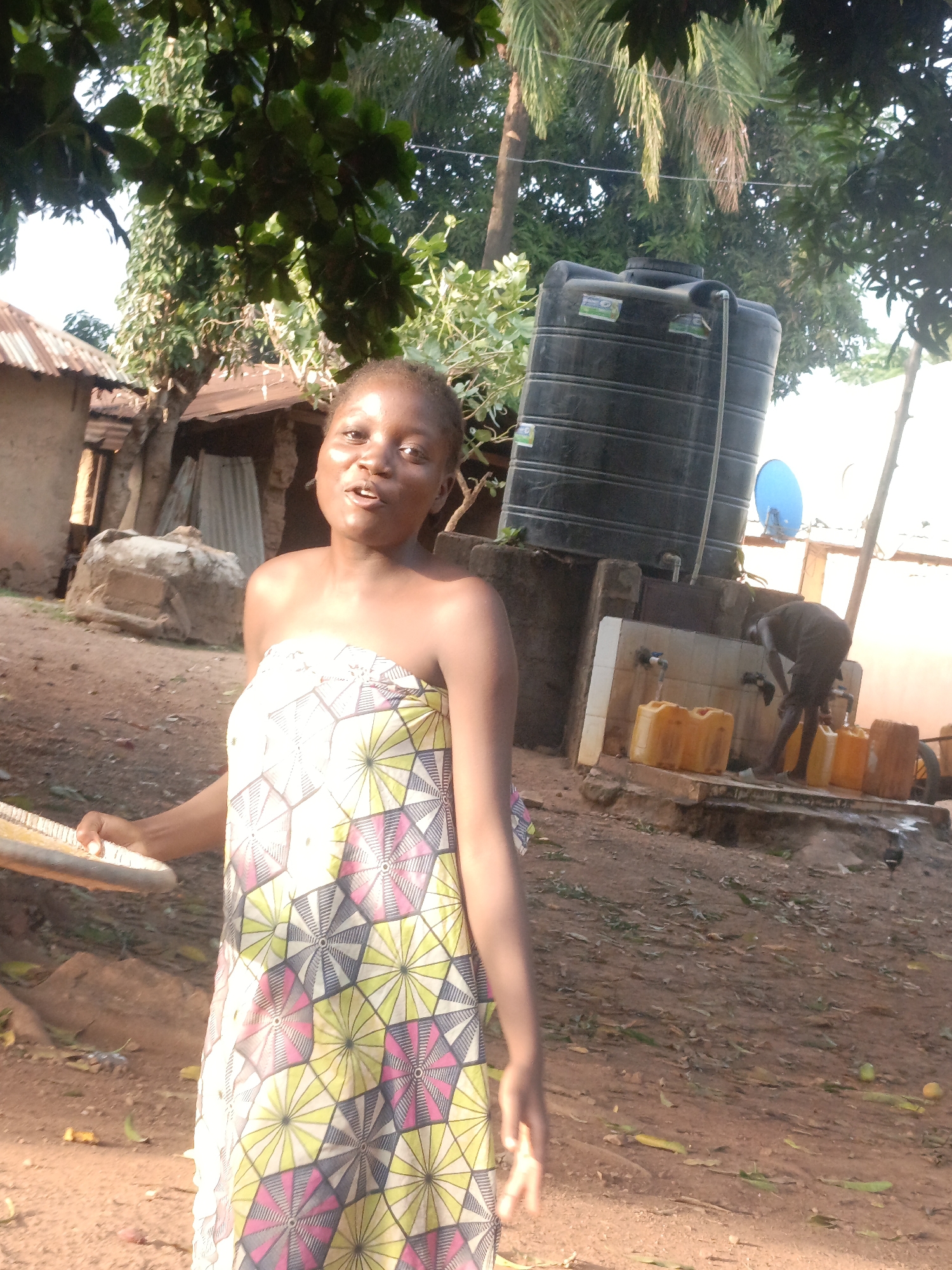 In Akwanga, Nigeria, a woman wearing a patterned yellow, white, and pink wraparound garment stands in a yard, holding a platter. Behind her, a black water tank is visible next to a concrete structure with faucets. A man is using the faucets to fill yellow jerrycans. The ground is dirt with scattered leaves and debris. In the background are structures appearing to be simple dwellings. Overhead, trees cast shadows on the scene.