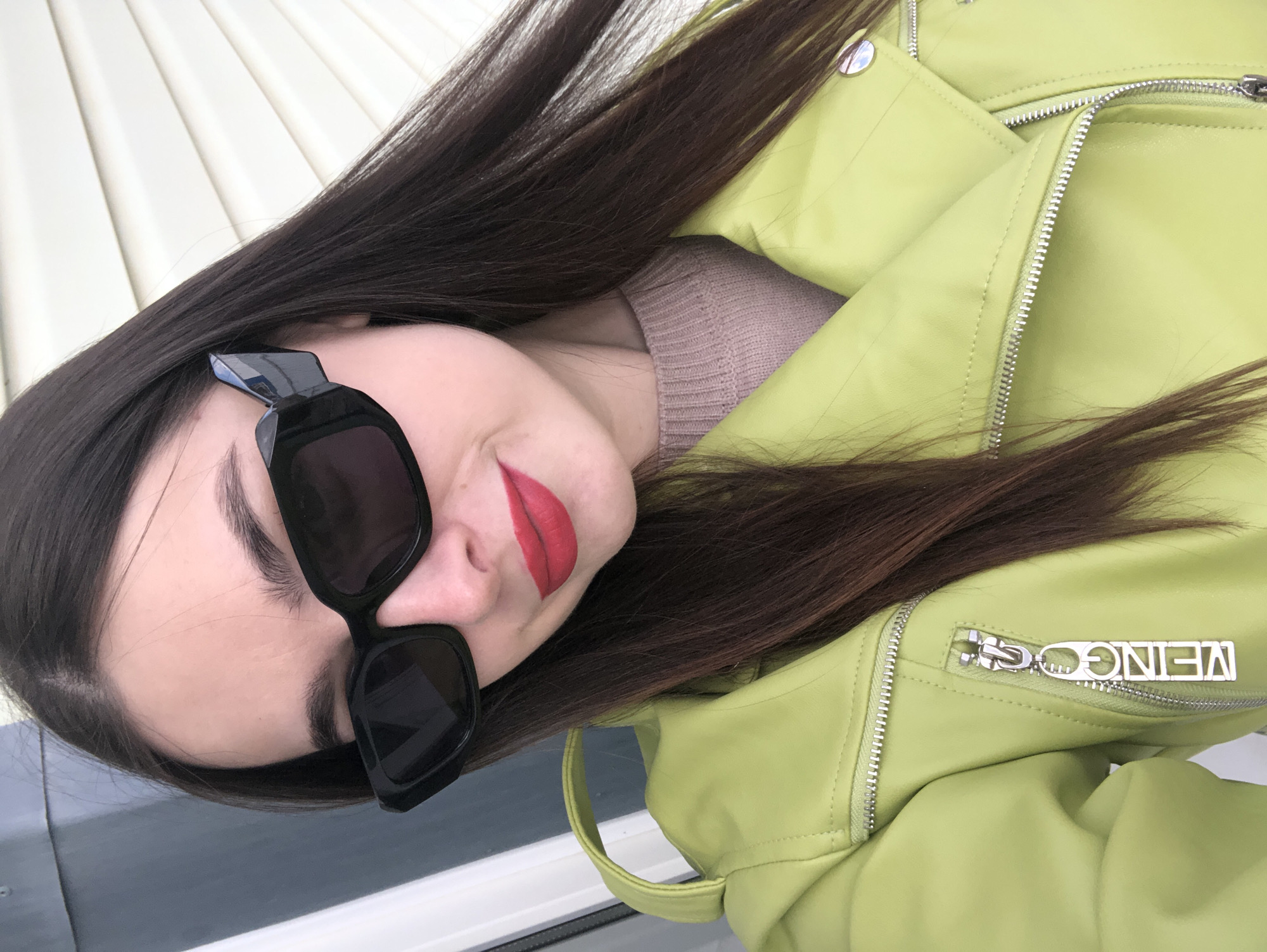 A close-up, portrait-style image captures a young woman from a slightly high angle with her head tilted. She has long, dark brown hair that cascades around her face and over her shoulders. Her eyes are obscured by large, black, square-framed sunglasses, but a subtle, closed-mouth smile is visible on her lips, which are painted with bright red lipstick.
She is dressed in a light green, possibly faux leather, biker-style jacket featuring prominent silver zippers and snap buttons on the lapels. One zipper is partially open, revealing a ribbed, light pink or beige knitted top worn underneath. A silver zipper pull on the jacket's chest pocket displays the word "VENCER" in block letters, appearing inverted due to the image's orientation.
The background is softly focused, showing a white, horizontally paneled surface, possibly building siding or blinds, on the upper left, and a darker, somewhat reflective surface, perhaps a window or railing, on the lower right. The scene is well-lit, indicating daytime. The photograph presents a casual, fashionable moment in Noviy, Russia.