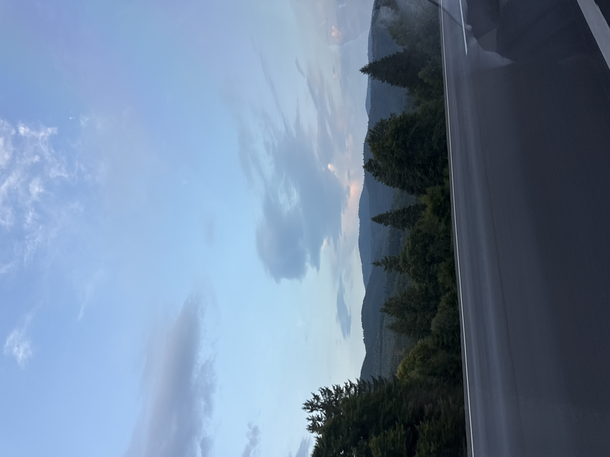 The image shows a view from a road overlooking a forested mountain range at dusk. The sky is a pale blue with scattered clouds, some of which are tinged with pink. The mountains in the background are dark and silhouetted against the sky. In the foreground, there is a dark asphalt road with a white line marking the lane. On the left side of the road, there are tall evergreen trees. The overall impression is one of peaceful, natural beauty. The location is the city of Brețcu, Romania.