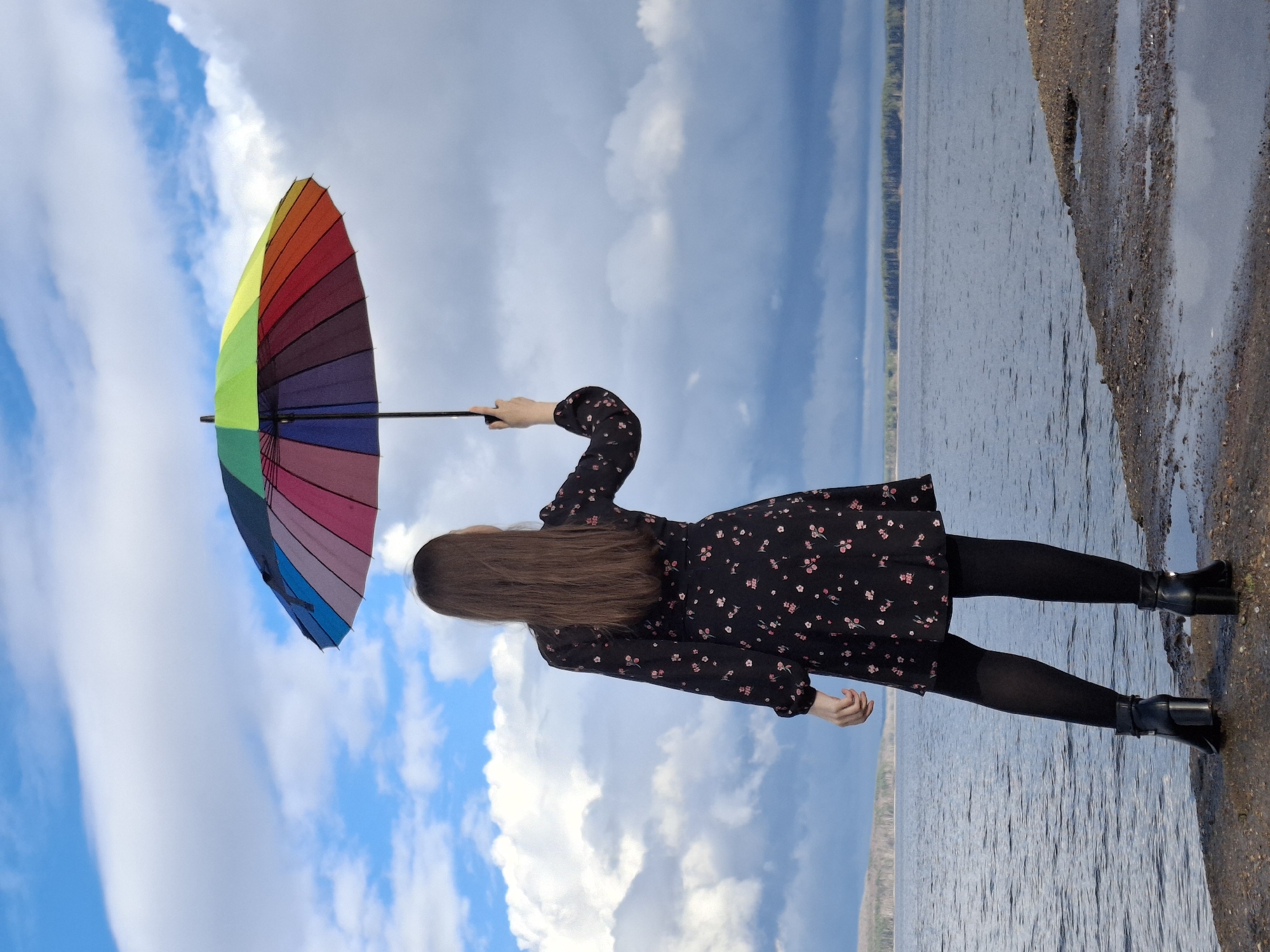 A woman with long brown hair stands on a wet, sandy shore holding a rainbow-colored umbrella. She is wearing a black floral dress, black tights, and black boots. Behind her is a body of water and a sky filled with blue and white clouds. The photograph was taken in the city of Chaikovsky, Russia.