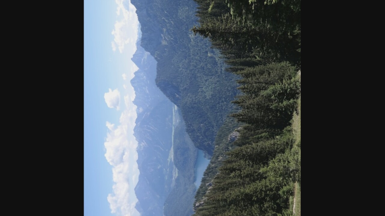 The image depicts a scenic mountain landscape with a clear blue sky and scattered white clouds. In the foreground, dense green pine trees cover the slopes of hills. Beyond these, a serene blue lake is visible, nestled within a valley. Further in the background, layers of progressively paler, misty mountains stretch towards the horizon. The overall scene is one of natural beauty and tranquility, characteristic of a mountainous region. The location is not specified as Oreshki, Russia, and there are no people, subjects, objects, or actions mentioned in the image.