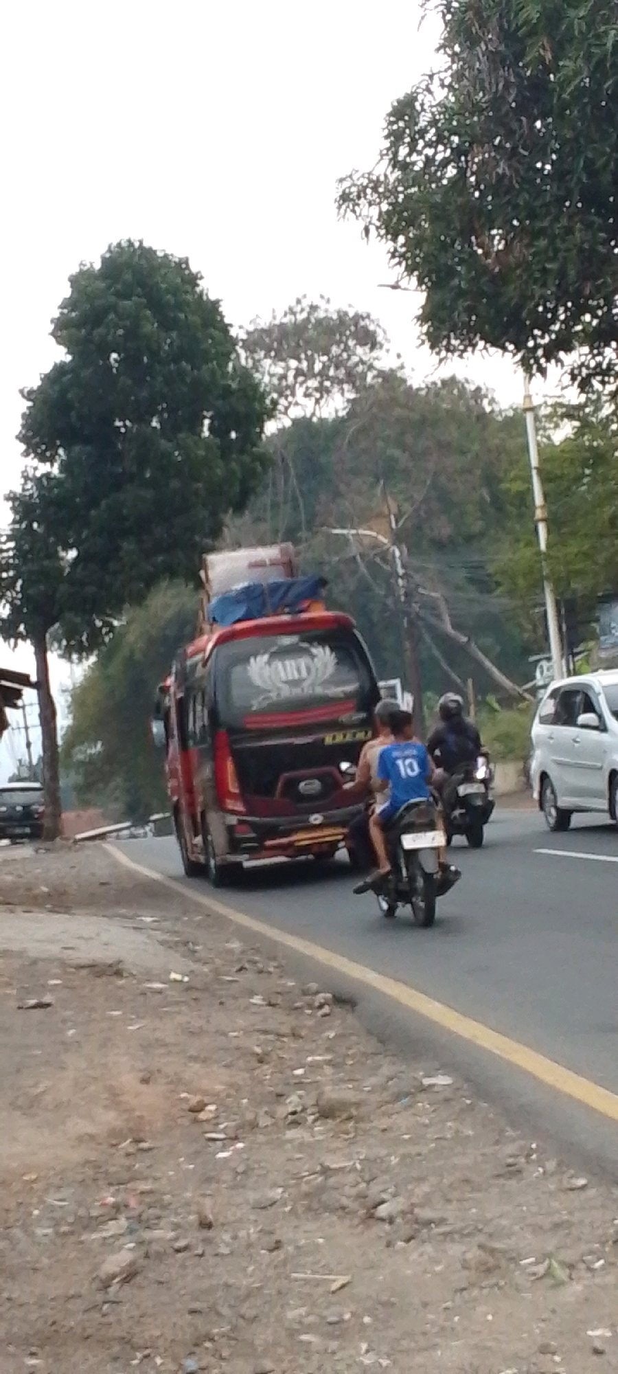 The image depicts a street scene in Cilaku, Indonesia, likely during daytime under an overcast sky. A red and black van is stopped on the right side of the road, its rear end facing the camera. The van has luggage strapped to its roof and a logo on its back window that appears to read "ART". To the right of the van, a white car is parked, partially visible.
In the foreground, a motorcycle carrying two individuals is traveling away from the camera on the paved road. The rider is wearing a dark helmet, and the passenger, a younger male, is wearing a blue t-shirt with the number "10" on the back. The road has a yellow dividing line.
The environment consists of a dirt shoulder on the left side of the road, littered with debris. Beyond the road, lush green trees and vegetation form a dense backdrop, with a utility pole visible on the far right. The overall impression is a rural or semi-urban road in Indonesia with typical local traffic.