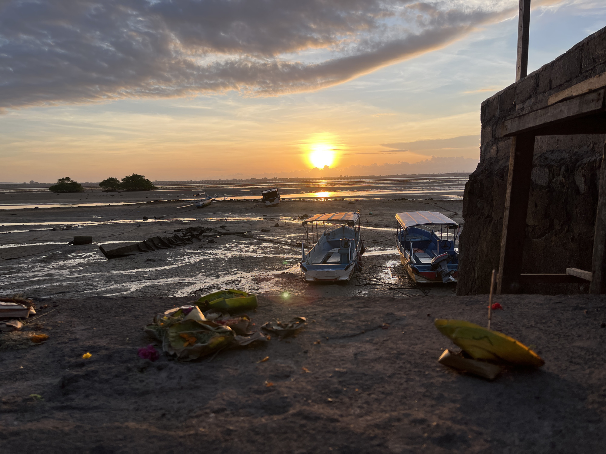 The image captures a sunset scene in Kuta Selatan, Indonesia. The sun is setting over a mudflat with several boats resting on the exposed seabed. In the foreground, there are offerings consisting of leaves, flowers, and food placed on a concrete surface. The boats are small with canopies. To the right, a structure partially frames the scene. The tide is low, revealing the mudflats and patches of water reflecting the sunset. The sky displays a mix of colors, with orange and yellow hues from the setting sun, and gray clouds.