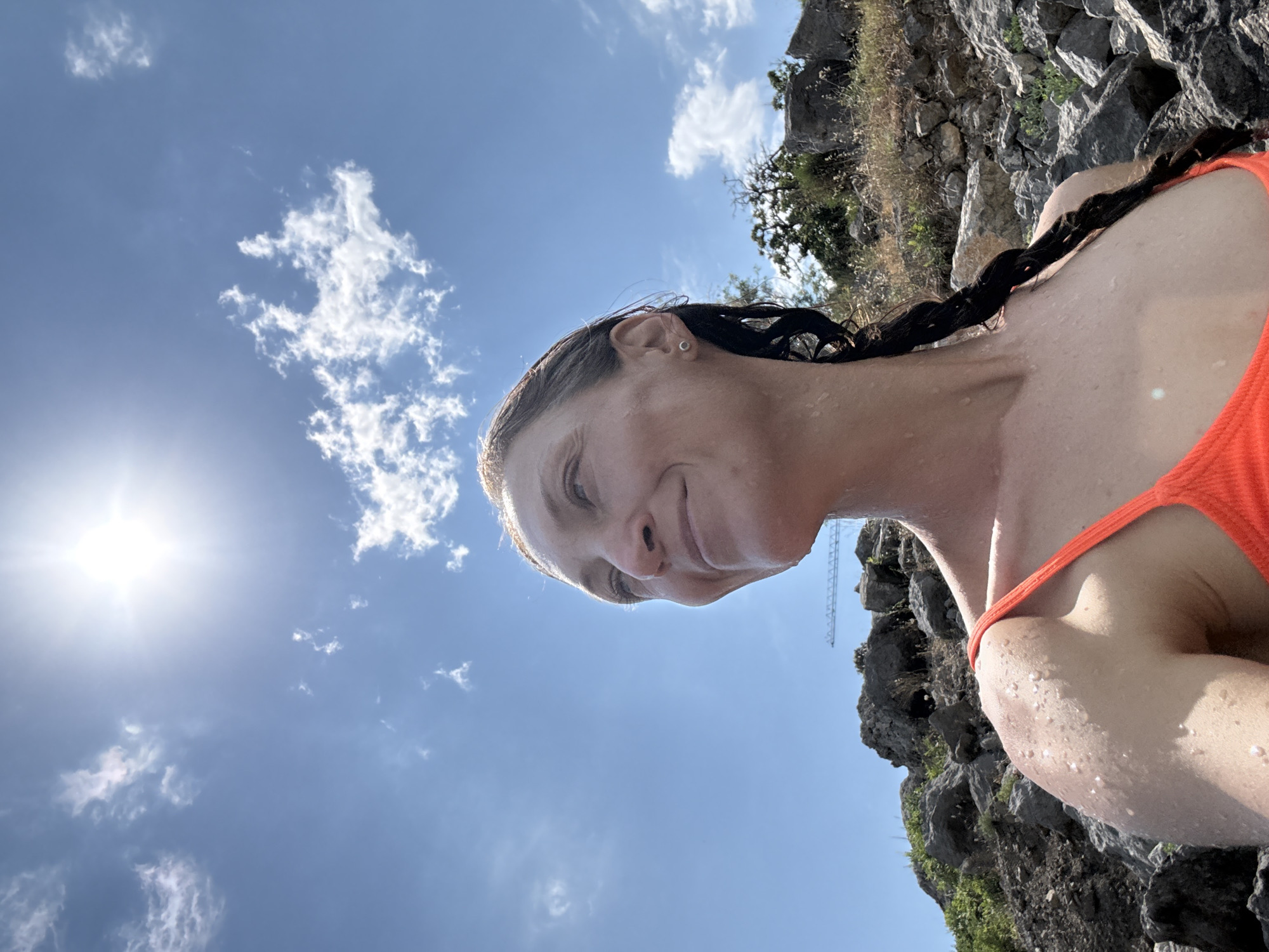 A selfie captures a woman in an orange bikini, her face tilted upwards and slightly to the left, with a gentle smile. Her hair, dark and wet, is braided down her back, and water droplets glisten on her skin, suggesting she has recently been in the water. The background consists of a rocky, sun-drenched landscape under a bright blue sky with scattered white clouds. In the distance, a construction crane is visible on the rocky terrain. The sun is high in the sky, creating a lens flare effect. The scene appears to be outdoors in a natural, possibly coastal or mountainous, environment. The location is specified as Bondarenkovo, Ukraine.