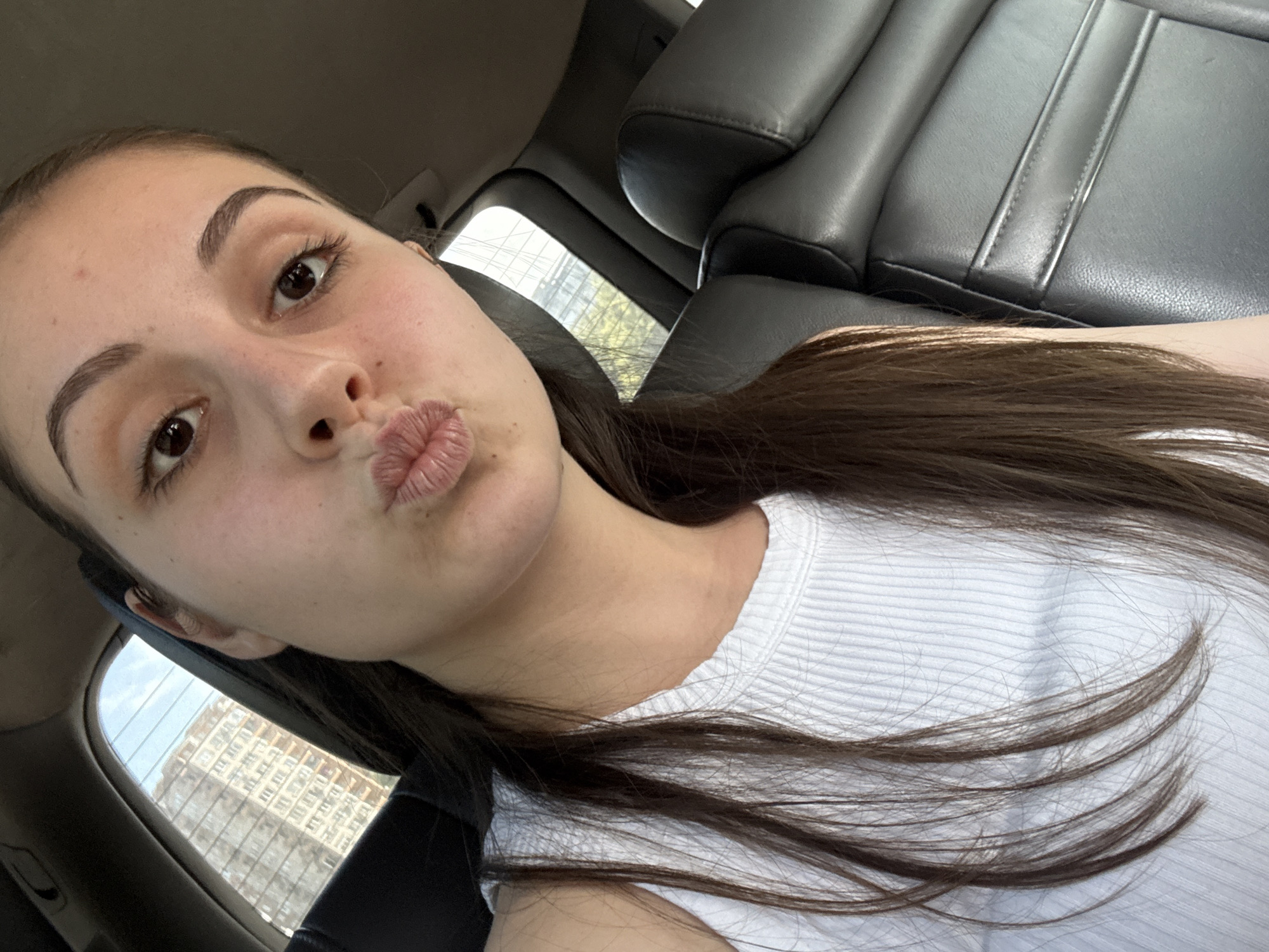 A selfie captures a young woman from a slightly upward angle inside what appears to be a car. Her face, positioned centrally, features dark, well-defined eyebrows, brown eyes, and lips pursed in a "kissing" expression. Her long, dark brown hair cascades over her right shoulder and chest, partially covering her white, ribbed top.
In the background to the right, dark grey or black leather car seats are visible, suggesting the interior of a vehicle. Through the car windows on the left and in the distance behind her, natural daylight streams in, illuminating the scene. A multi-story building with many windows can be distinctly seen through one of the windows, indicating an urban or suburban environment. The clear view outside suggests fair weather conditions. The overall impression conveyed by her pose is playful. The image was taken in Burgas, Bulgaria. No text is visible in the media.