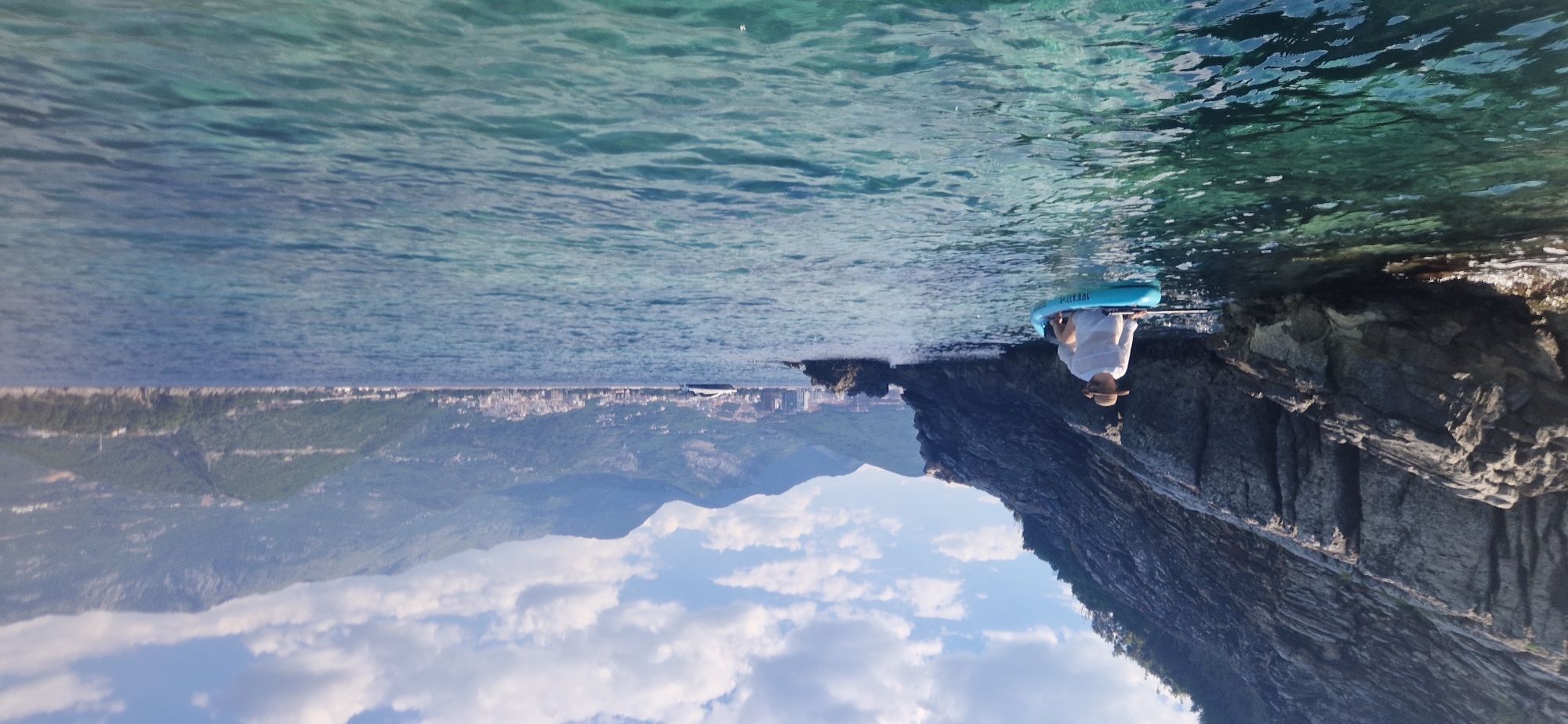 The image depicts a serene scene at Boreti, Montenegro, with a person on a paddleboard near a rocky shoreline. The water is clear and turquoise, revealing the sandy bottom beneath. The sun appears to be shining, suggesting it's a bright day, possibly late morning or early afternoon given the lighting.
The main subject is a person dressed in white, wearing a straw hat, seated on a bright blue paddleboard. The person is holding a paddle, appearing relaxed and enjoying the peaceful environment. The paddleboard has visible text on it, though it is partially obscured. The shoreline is characterized by rugged, grey rocks that plunge into the water, creating a dramatic contrast with the calm sea.
In the background, a picturesque landscape unfolds. A coastal town with numerous buildings is visible, nestled against a backdrop of lush green hills that transition into majestic mountains under a partly cloudy sky. The clouds are white and fluffy, scattered across a vibrant blue sky, adding to the idyllic atmosphere. The orientation of the image is unusual, with the sky appearing below the water, suggesting the photograph may have been taken at an unconventional angle or has been rotated. The overall mood is tranquil and summery, evoking a sense of leisure and connection with nature.