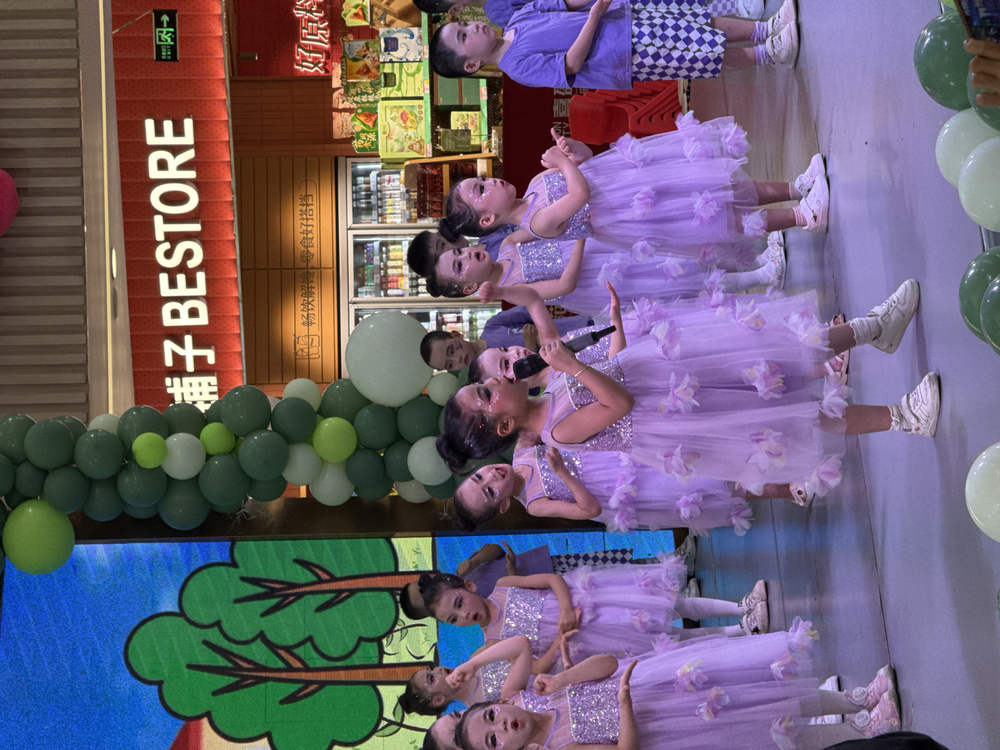 In Jiulongpo District, China, a group of children are performing in front of a Bestore store. The girls wear lavender dresses adorned with floral accents, while a boy is dressed in a purple shirt and checkered shorts. They stand on a stage decorated with green and white balloons, in front of a screen displaying a cartoon tree. Some of the children hold microphones and sing. The Bestore store is visible in the background, with refrigerators displaying food items.
