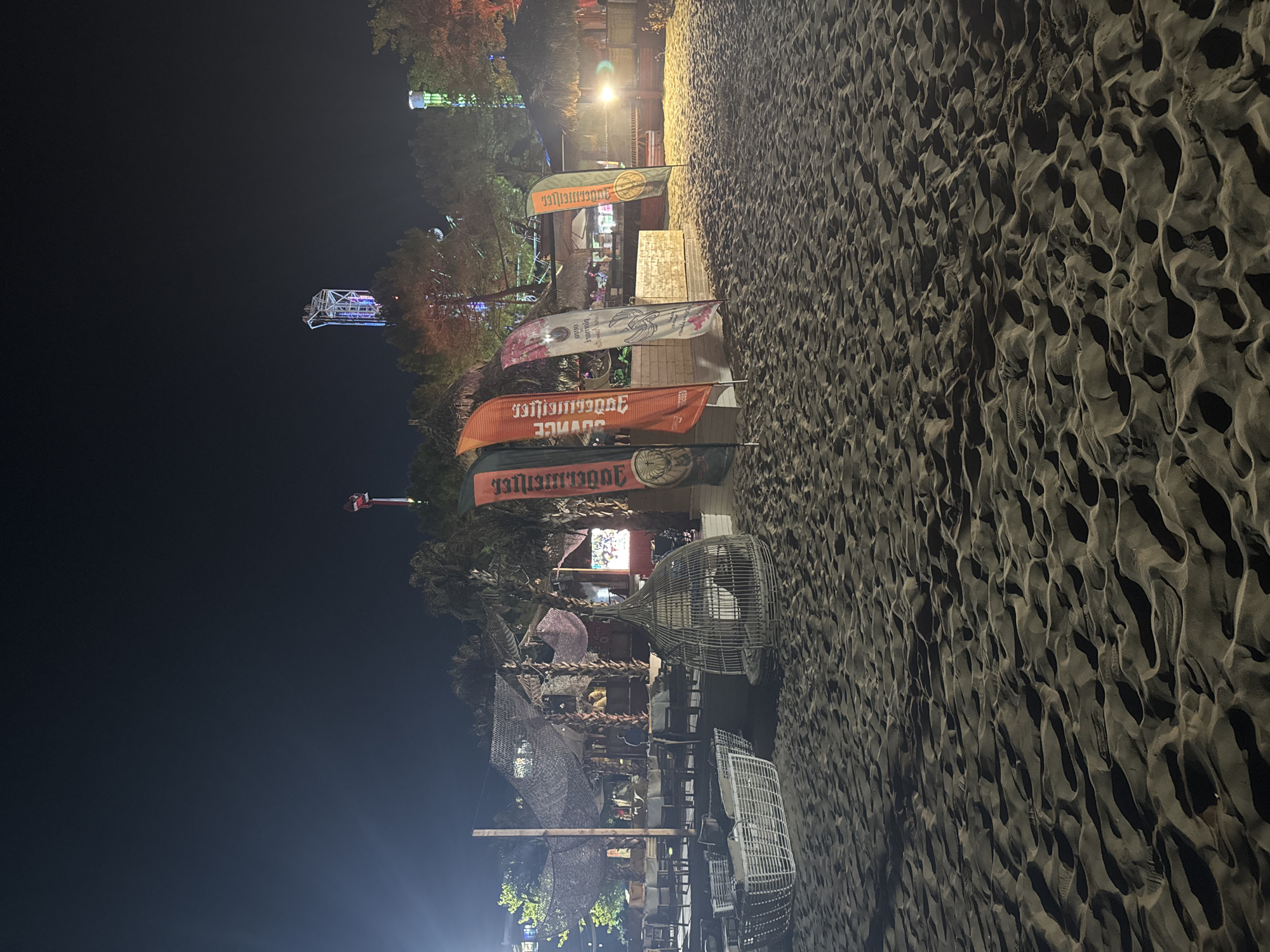 This is a nighttime outdoor photograph of a sandy beach in Nesebar, Bulgaria. The scene depicts a beach bar or club area with several flags displayed in front. The flags are vertical and appear to be advertising beverages, with one featuring the Jägermeister logo. Behind the flags, there is a rustic-looking structure with thatched roofs, illuminated by artificial lights. Several tables and chairs are set up in front of this structure, suggesting it is a seating area for patrons. In the foreground, two large, white wicker chairs or loungers are visible on the sand. Further in the background, amusement park rides, including a tall tower ride and a drop tower, are illuminated and visible against the dark night sky. The overall atmosphere is that of a lively nighttime entertainment venue on a beach.