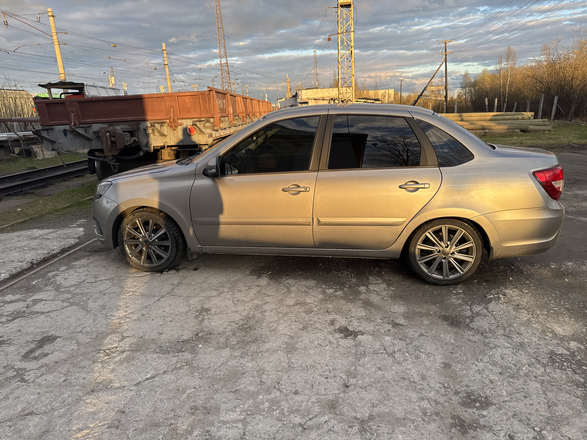 The image depicts a silver Lada Granta sedan parked on a cracked concrete surface. The location appears to be an industrial area in Apatity, Russia. In the background, there are railway tracks and a freight wagon, along with utility poles and power lines. A pile of logs is visible in the distance. The sky is overcast with a mix of clouds and sunlight. There are no people visible in the photograph.