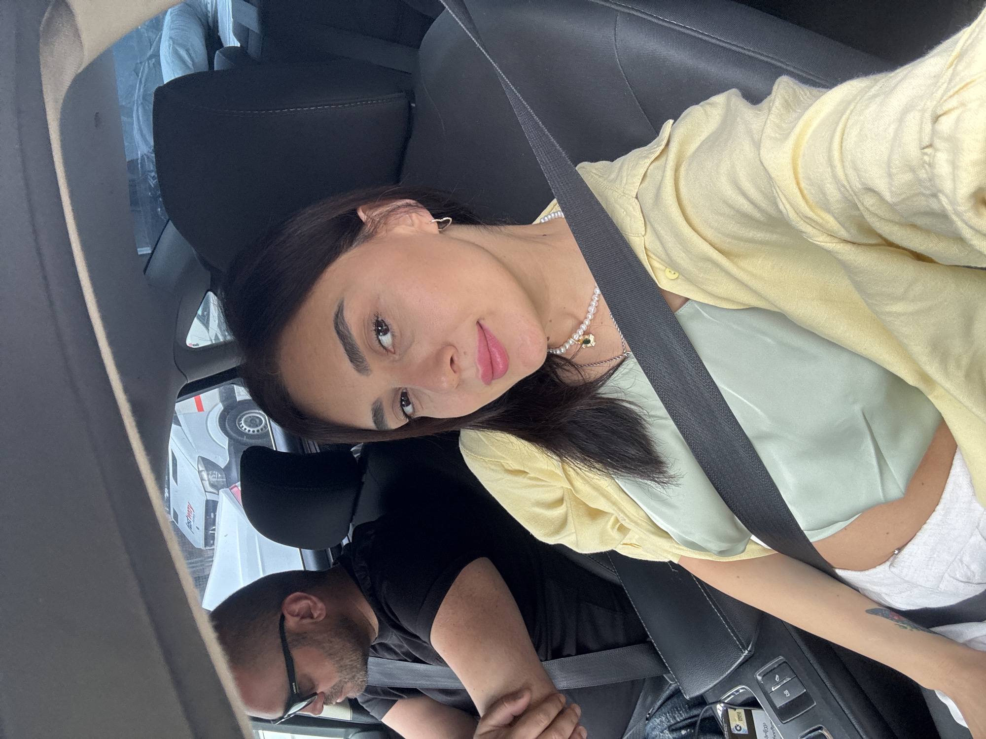 The image shows a woman and a man sitting in a car. The woman is in the foreground, taking a selfie. She has long dark hair, brown eyes, and is wearing a yellow button-up shirt over a light green top and white pants. A pearl necklace with a small gold pendant is visible around her neck. A seatbelt crosses diagonally across her body.
The man is in the background, seated in the driver's or passenger's seat. He is wearing glasses and a black t-shirt. His arm is resting on something, and he appears to be looking forward.
The scene appears to be an interior of a car. Outside the car, through the windows, another vehicle, possibly a white truck with some red lettering, is visible. The image is taken in daylight. The overall impression is that the two individuals are traveling together. The location is identified as Tbilisi, Georgia. There is some text visible on a card or sticker in the car's center console, but it is not legible.