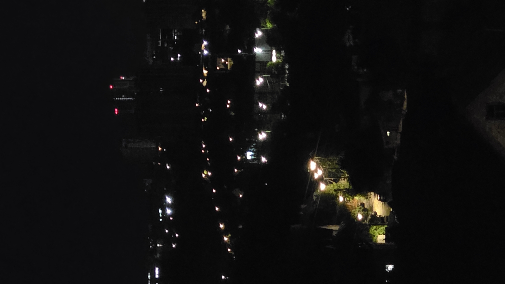A vertical, nighttime, long-range photograph captures a sprawling cityscape illuminated by numerous artificial lights against a black sky. The composition is dominated by the darkness of night, punctuated by the glow of lights that reveal a complex urban or suburban landscape. In the far distance, silhouetted against the sky, are several tall buildings. Two of these buildings are particularly prominent, each topped with red lights, suggesting they are either communication towers or buildings with navigational lighting.
Closer to the foreground, a dense collection of lights indicates a neighborhood with residential or commercial structures. Streetlights, appearing as small, bright points, are scattered throughout the mid-ground, lining what seem to be roads or pathways. Some of these lights are yellow or white, while others have a slight pinkish hue, possibly due to their type or distance.
In the lower portion of the image, the lights become more concentrated, illuminating sections of what appear to be homes or businesses nestled amongst dark foliage. A cluster of lights, arranged in a string, hangs over a pathway or garden area, possibly from decorative string lights. One building in the lower right quadrant of the image has a dimly lit window, providing a faint square of light. The overall impression is of a city or town at night, viewed from a high vantage point, with a clear contrast between the illuminated structures and the pervasive darkness of the night.