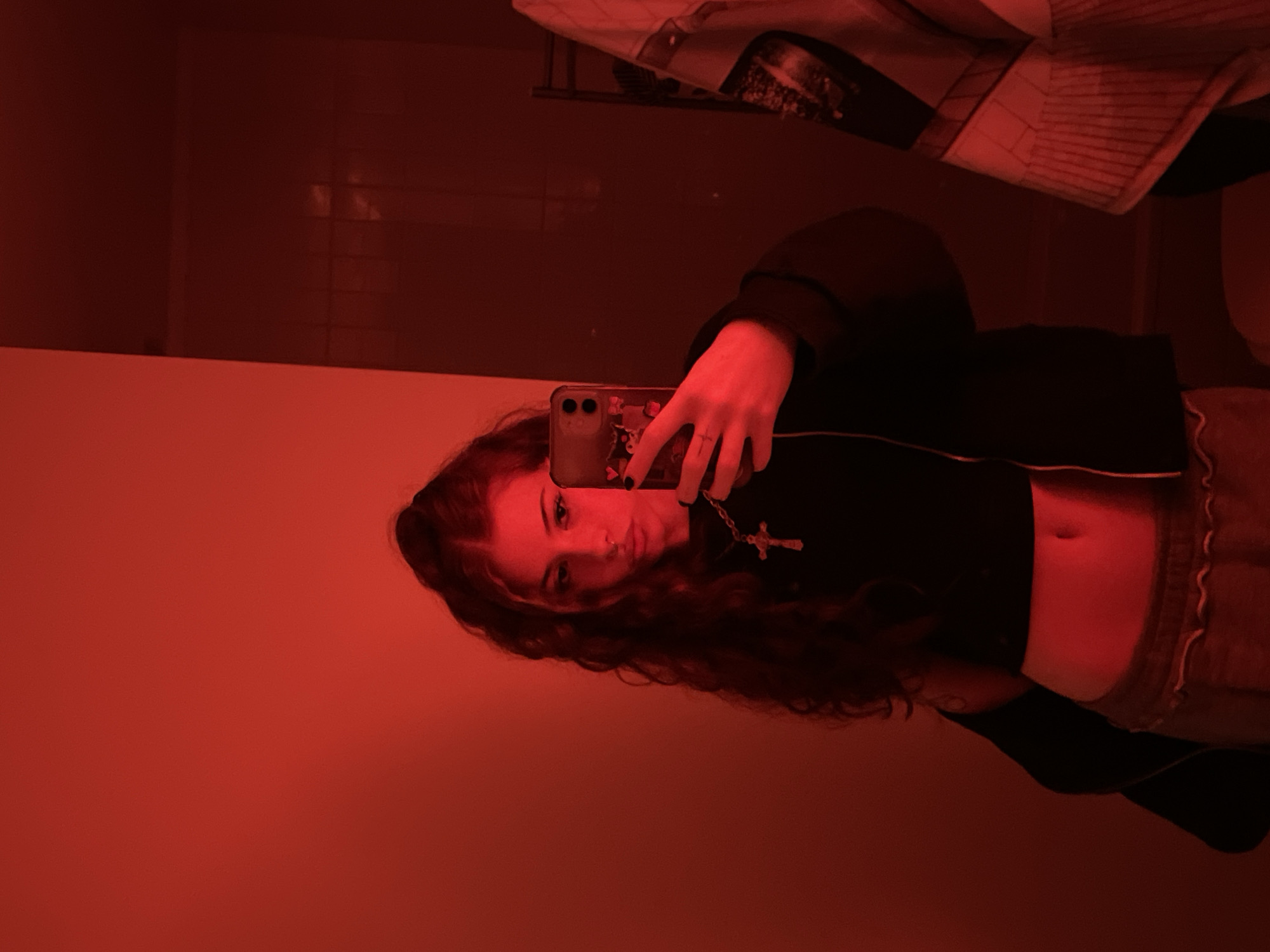 The scene depicts a young female individual in an interior space, bathed entirely in intense red lighting. She is positioned slightly left of center, visible from the mid-torso upwards, and appears to be taking a mirror selfie or a direct camera shot. She possesses long, dark, wavy hair extending over her shoulders and wears a small ring piercing in her left nostril. Her attire includes a dark, cropped top exposing her midriff and light-colored bottoms with a visible ruffled waistband. A necklace with a prominent silver-colored cross pendant is worn around her neck. Her left hand (viewer's right) holds a smartphone with a clear case adorned with numerous red heart graphics; her fingernails are painted dark. The background to the left is a plain red wall. To the right, a darker vertical surface, possibly a wall or door, is visible, partially obscuring a metallic rack structure. A textile item with a white and red geometric pattern, including stripes and rectangular shapes, along with a darker abstract graphic, hangs from this rack. The exact location within the city of Buenos Aires, Argentina, cannot be determined from the visual information provided.