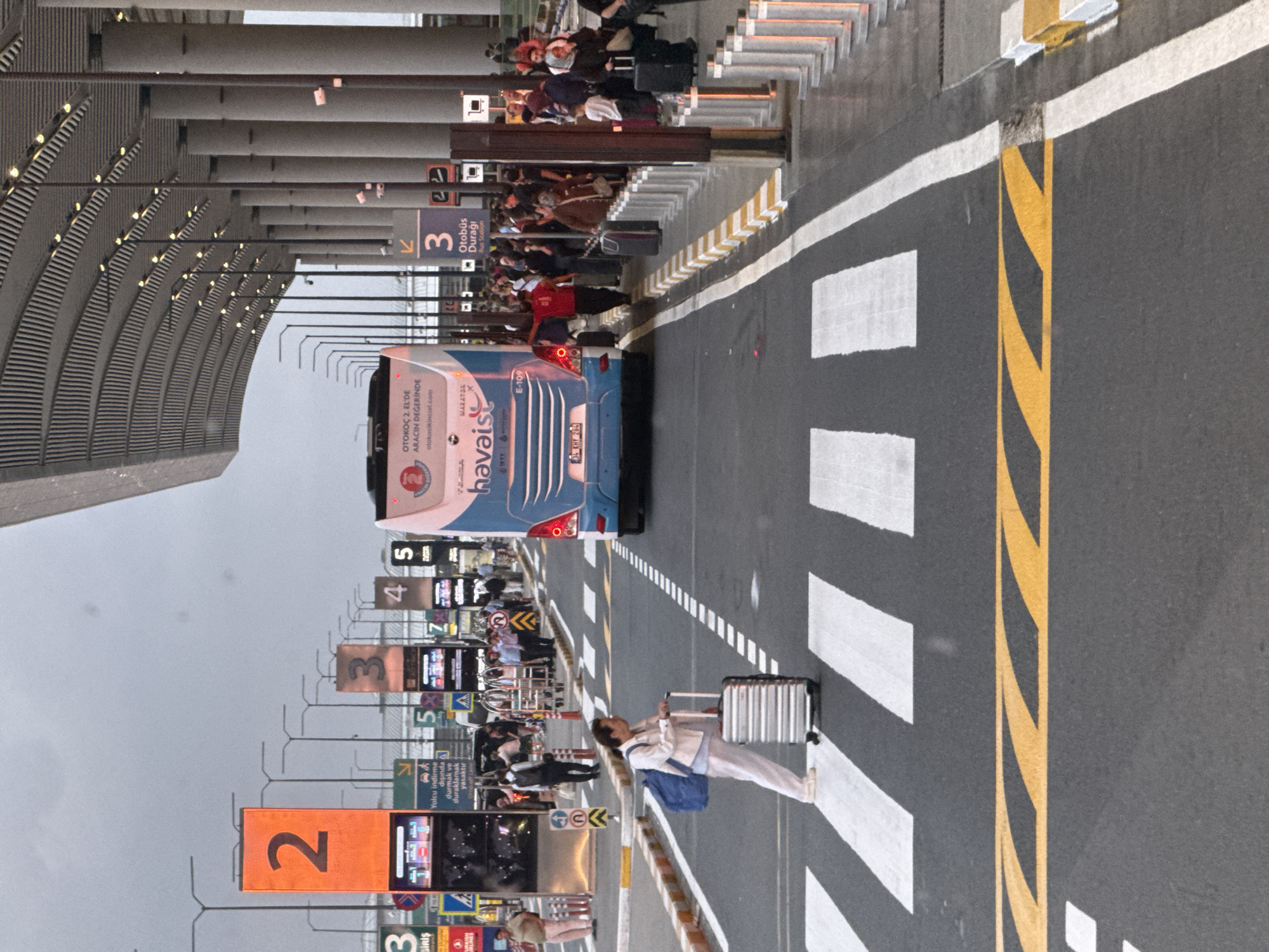 The image depicts the exterior of a modern airport terminal in Arnavutköy, Turkey, likely Istanbul Airport given the presence of the "HAVAİST" bus service. A large, blue and white bus, identified as a HAVAİST bus with license plate "34 KHY 286", is positioned on the roadway, partially obscuring a crosswalk. Several people are visible near the bus station and on the sidewalk, some with luggage, suggesting transit activity. One woman, dressed in a light-colored jacket and pants, is crossing the crosswalk with a rolling suitcase and a blue backpack. In the background, tall pillars support a distinctive, undulating roof structure characteristic of airport architecture. Various signage is present, including large orange and black numbers "2", "3", "4", and "5", indicating bus bays or zones, as well as other informational signs in Turkish. The ground is paved with asphalt, marked with white and yellow pedestrian crosswalks and directional lines. The sky is overcast, indicating a cloudy day. The overall scene captures a moment of passenger movement and transportation infrastructure at an airport.