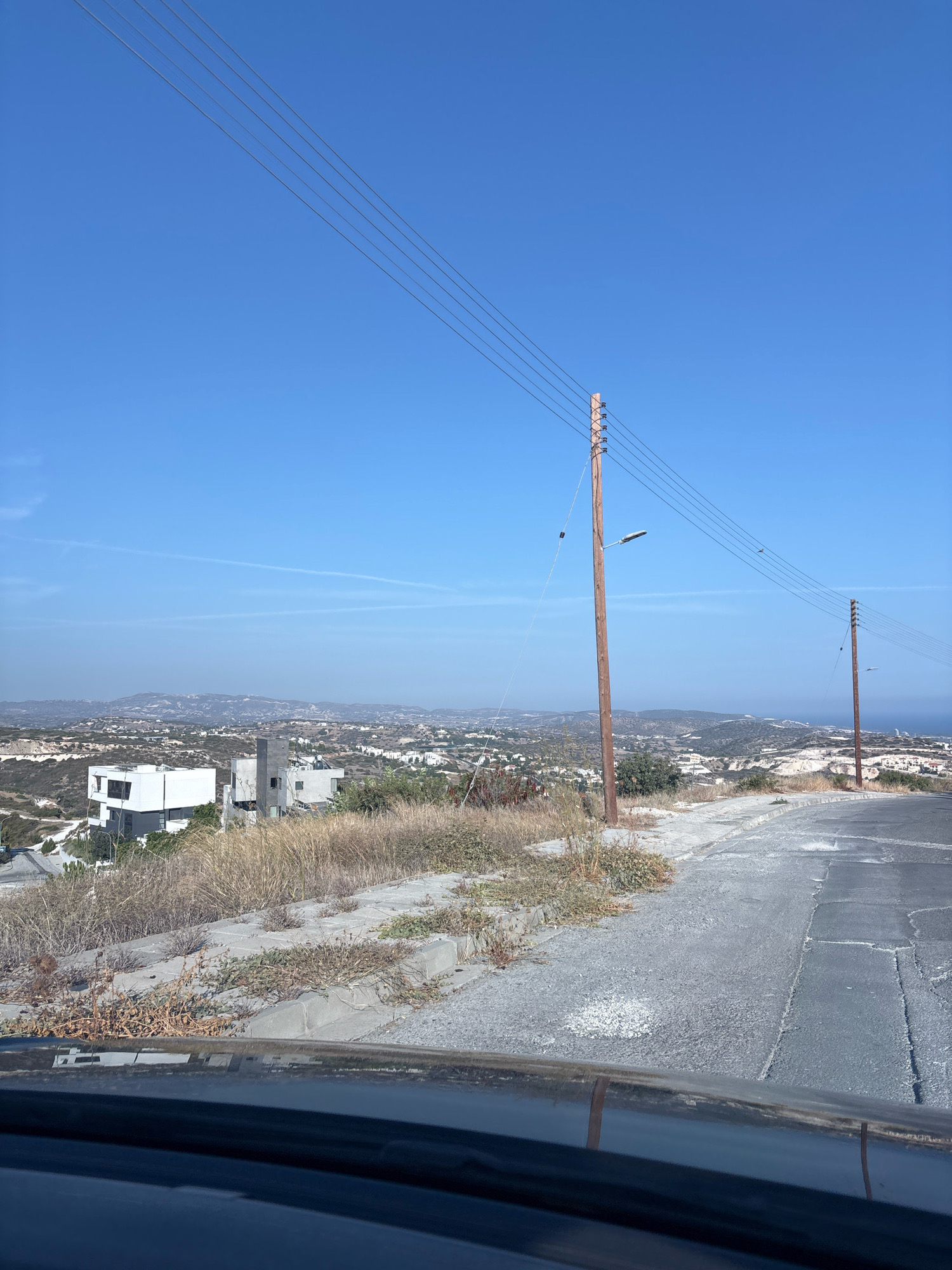A daytime, medium shot, captured from a vehicle's perspective, shows a road curving right through a dry, hilly landscape in Agios Tychona Community, Cyprus. The road, paved with asphalt and exhibiting some wear and tear, is flanked by a concrete curb and dry, sparse vegetation. In the middle ground, a cluster of modern, geometric white buildings with dark accents is visible, some appearing partially constructed. Further in the distance, a sprawling town or village stretches across undulating hills, with the sea visible in the far distance under a clear, bright blue sky. Two wooden utility poles with multiple wires and insulators stand prominently along the roadside. One pole, closer to the viewer, also features a streetlamp. The overall impression is one of arid, developing terrain with a coastal view.