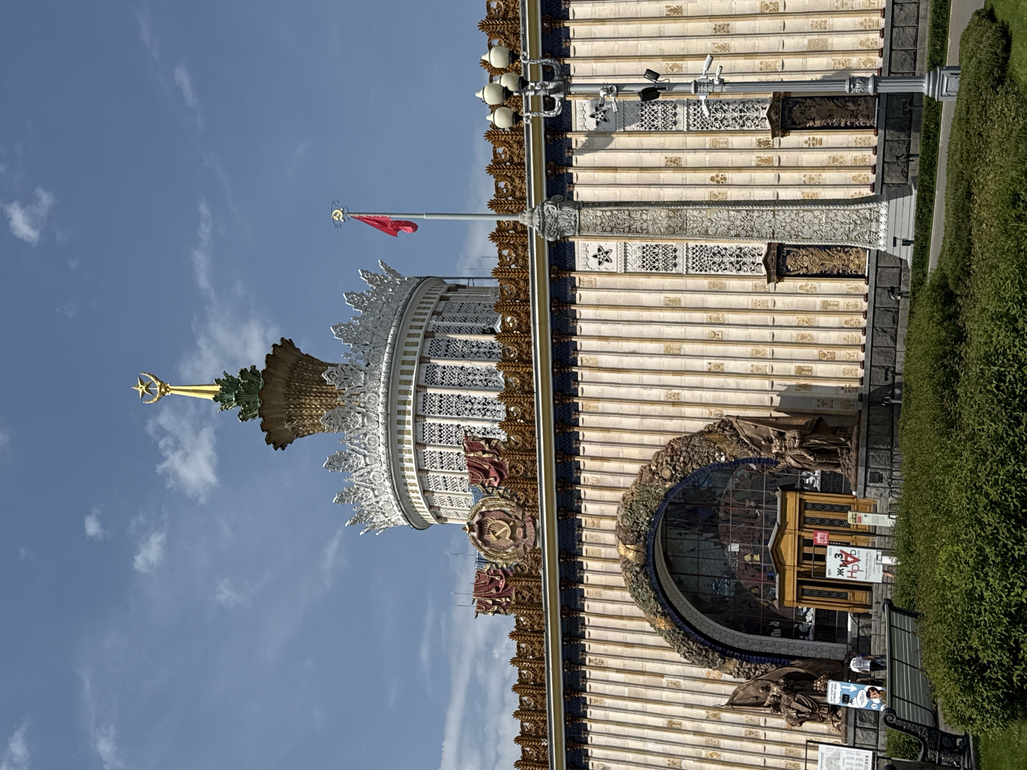 The image shows a section of the Pavilion №1 "Central" at VDNKh, a permanent general-purpose trade show and amusement park in Moscow, Russia. The pavilion features a large arched window entrance framed with detailed sculptures. The building's facade is decorated with friezes and ornamentation. Atop the building is a tower structure adorned with intricate white and gold details, topped with a golden star. A red flag flies nearby. The sky is partially cloudy. Green bushes line the base of the building.
