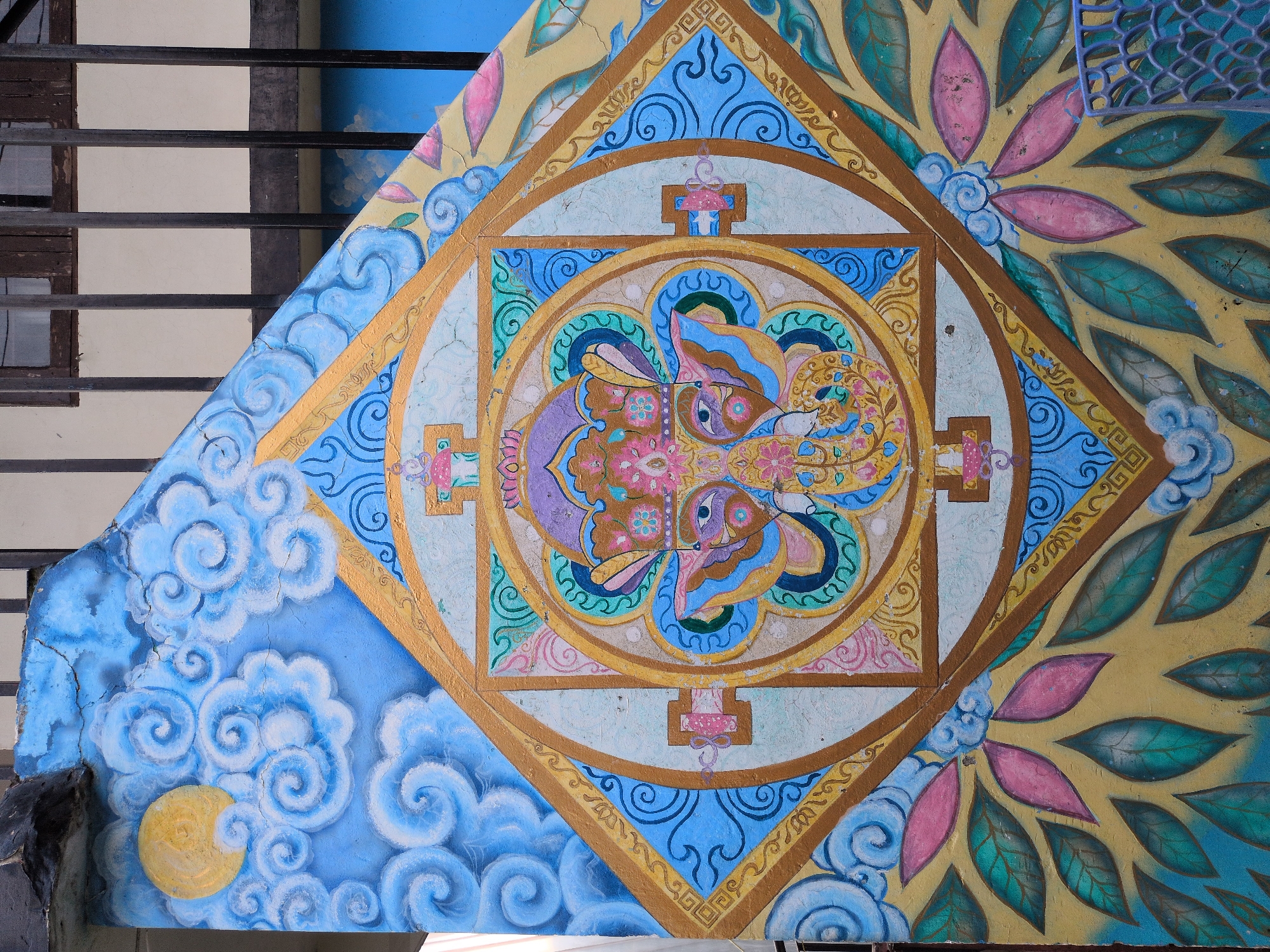 The image displays a colorful mural painted on an exterior wall, likely part of a building in Haini, India. The mural is intricately detailed, featuring a central mandala-like design with an elephant head motif. Surrounding the central design are swirling clouds painted in shades of blue and white, with a yellow full moon in the upper left quadrant. The lower portion of the mural depicts a pattern of large, stylized leaves in pink and teal against a yellow background. The mural is painted on a textured surface that appears to be aged, with some cracks visible. In the background, a dark metal fence and a portion of a building with windows are visible. The overall impression is one of vibrant religious or cultural art adorning an outdoor space.