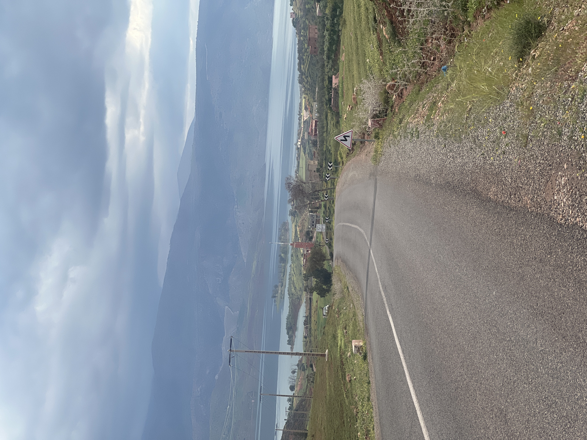 The image shows a scenic view of Bin El Ouidane, Morocco, captured from an elevated road. The road curves, revealing a lake surrounded by green hills and mountains. The sky is cloudy. Power lines are visible in the landscape. A winding road warning sign is present along the roadside. The landscape includes green vegetation and scattered buildings.