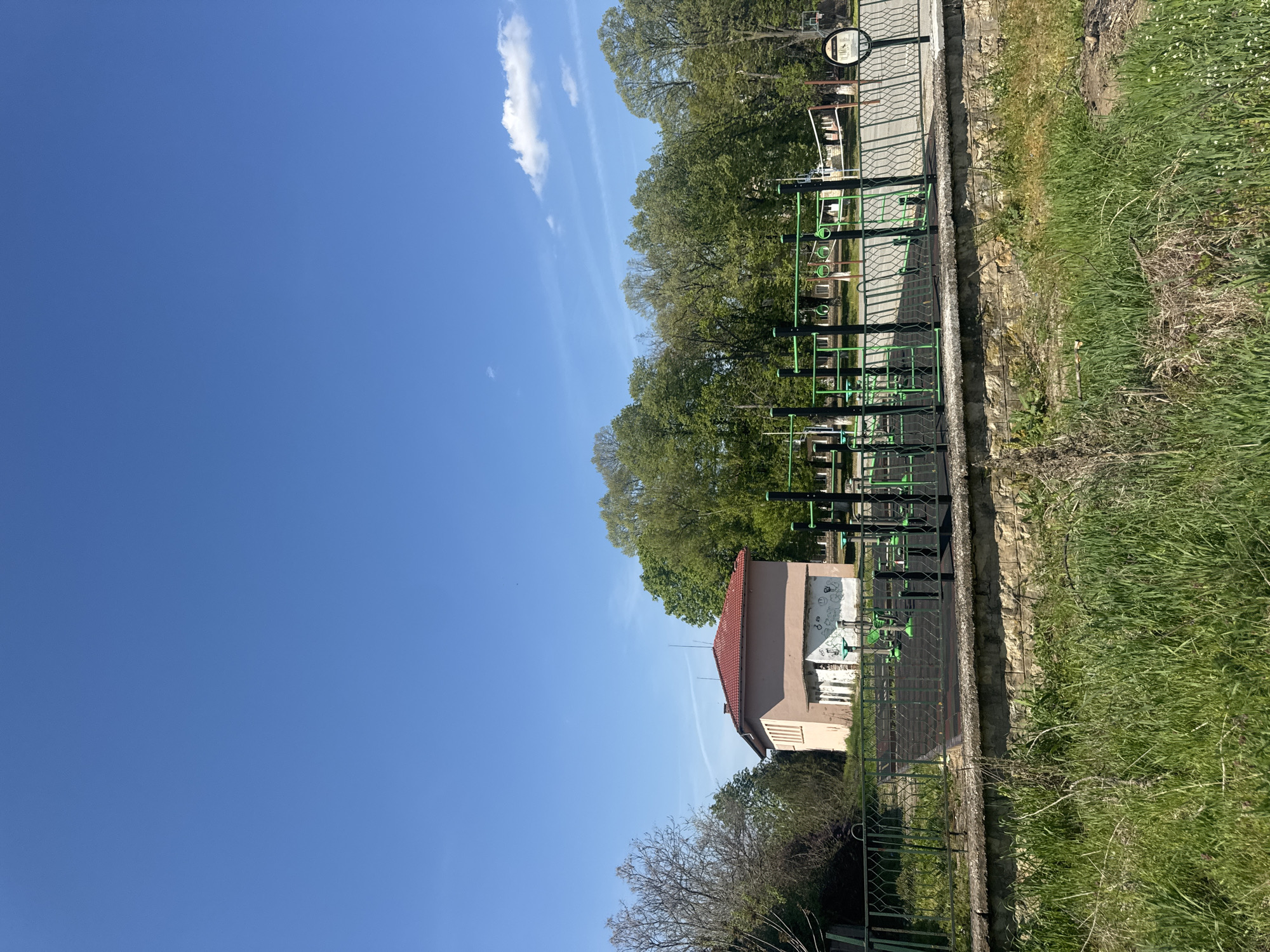 The image depicts an outdoor exercise park in Lehchevo, Bulgaria. The park features various green and black metal exercise structures. A small, two-story building with a red tile roof is adjacent to the park. Graffiti is visible on the side of the building. A fence runs along the perimeter of the park, separating it from an area of grass and vegetation. The sky is blue with some clouds.