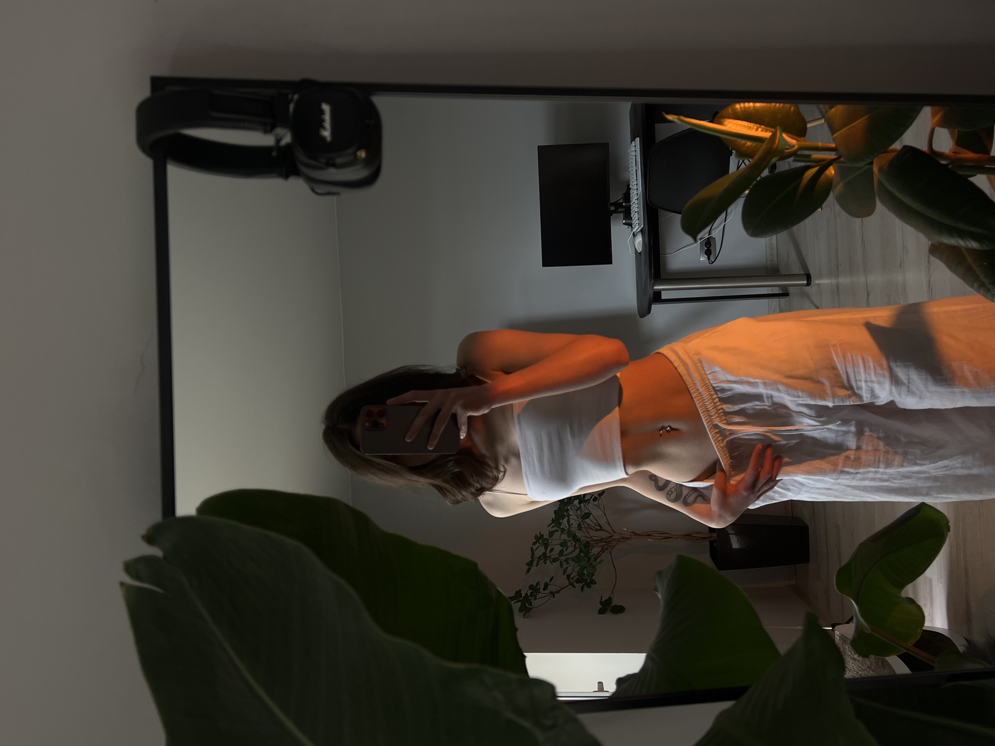 A person is taking a mirror selfie in a room. The person is wearing a white crop top and white pants. They have a tattoo on their left arm and a belly button piercing. In the background, there is a desk with a computer monitor, keyboard, and mouse, as well as a pair of headphones hanging on the mirror. There are also plants in the room. The lighting suggests it is either sunset or sunrise, casting an orange glow on the person. The location is not specified to be in Ternopil, Ukraine.
