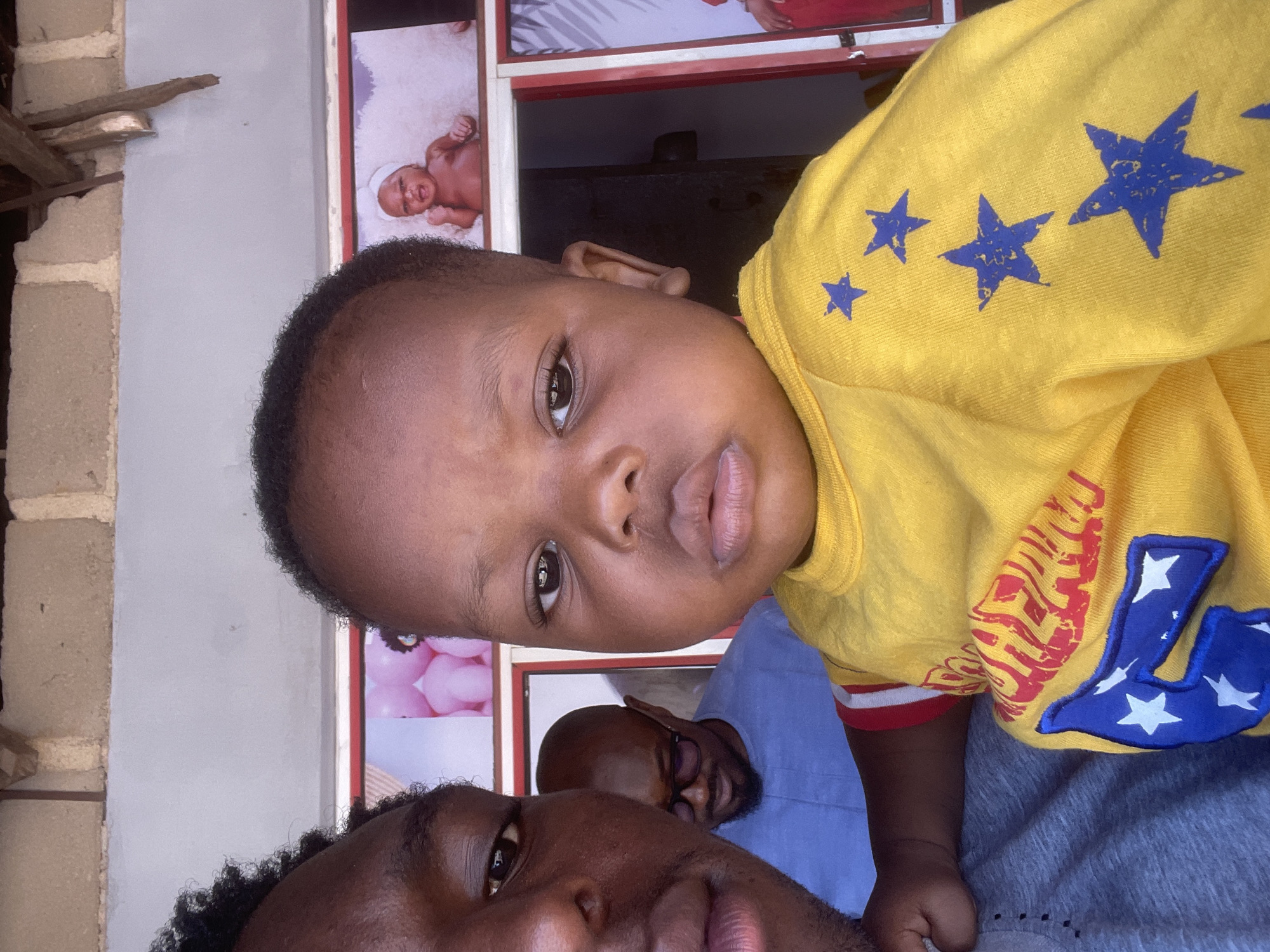 An outdoor selfie shows three individuals. The foreground is dominated by a young child in a yellow shirt with blue stars. To the left of the child, a person's face is partially visible, looking towards the camera. Behind them, a man wearing glasses and a blue shirt is looking down. In the background, there are framed pictures on a wall, including one of a baby. The scene appears to be in Barakin Akawo, Nigeria, with brickwork and a wooden structure visible overhead, suggesting an open-air setting.