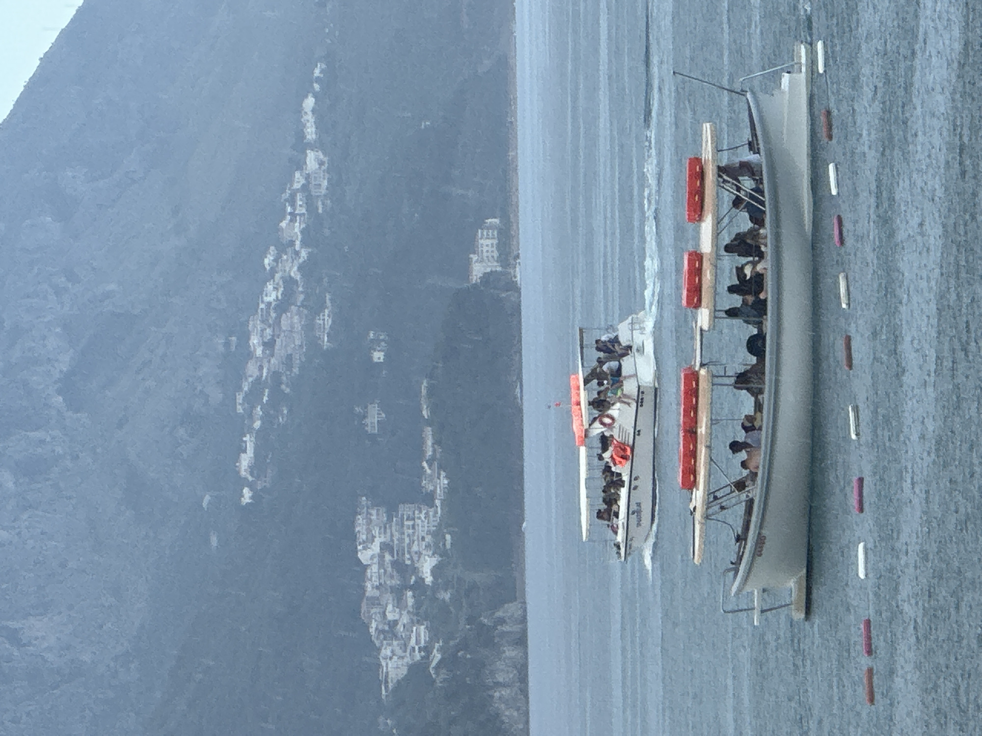 Two tour boats are on the water in front of a hillside covered in buildings. The boat in the foreground is white with a tan canopy and red life preservers on top. It has people sitting inside. The boat in the background is also white with a white canopy and orange life preservers on top. It is also full of people. The buildings on the hillside appear to be white and clustered together. The water is choppy and the sky is overcast. The scene is likely the coast of Boreti, Montenegro.
