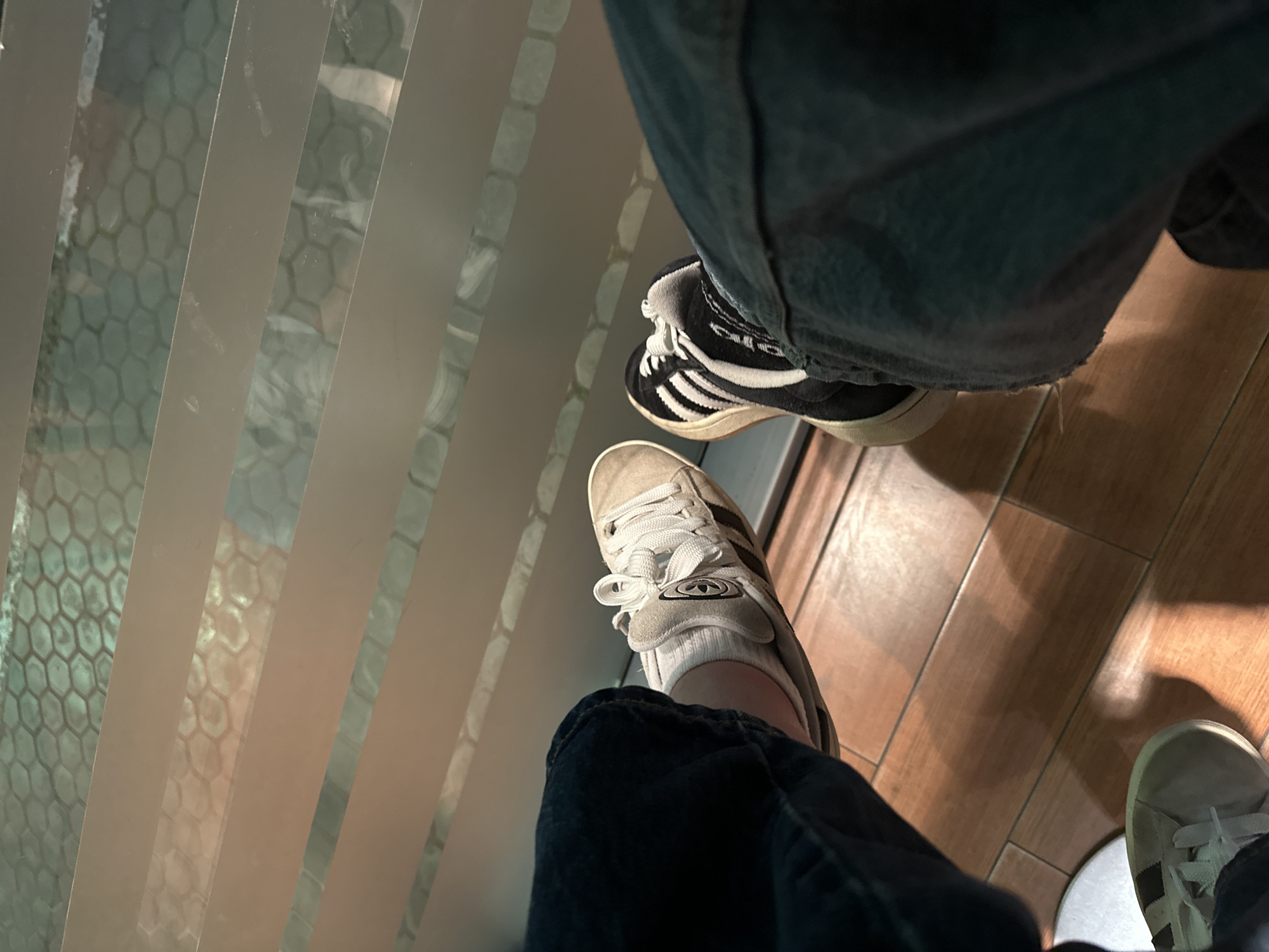 The image shows a first-person view of two pairs of Adidas Gazelle shoes being worn. The shoes are resting on what appears to be a glass surface with a geometric pattern. The person on the left is wearing beige Gazelles, and the person on the right is wearing black and white Gazelles. Both are wearing dark-colored jeans. Based on the background visible through the glass, it is difficult to pinpoint the exact location in Stara Zagora, Bulgaria. More information about the environment seen through the glass would be needed for a specific location identification.