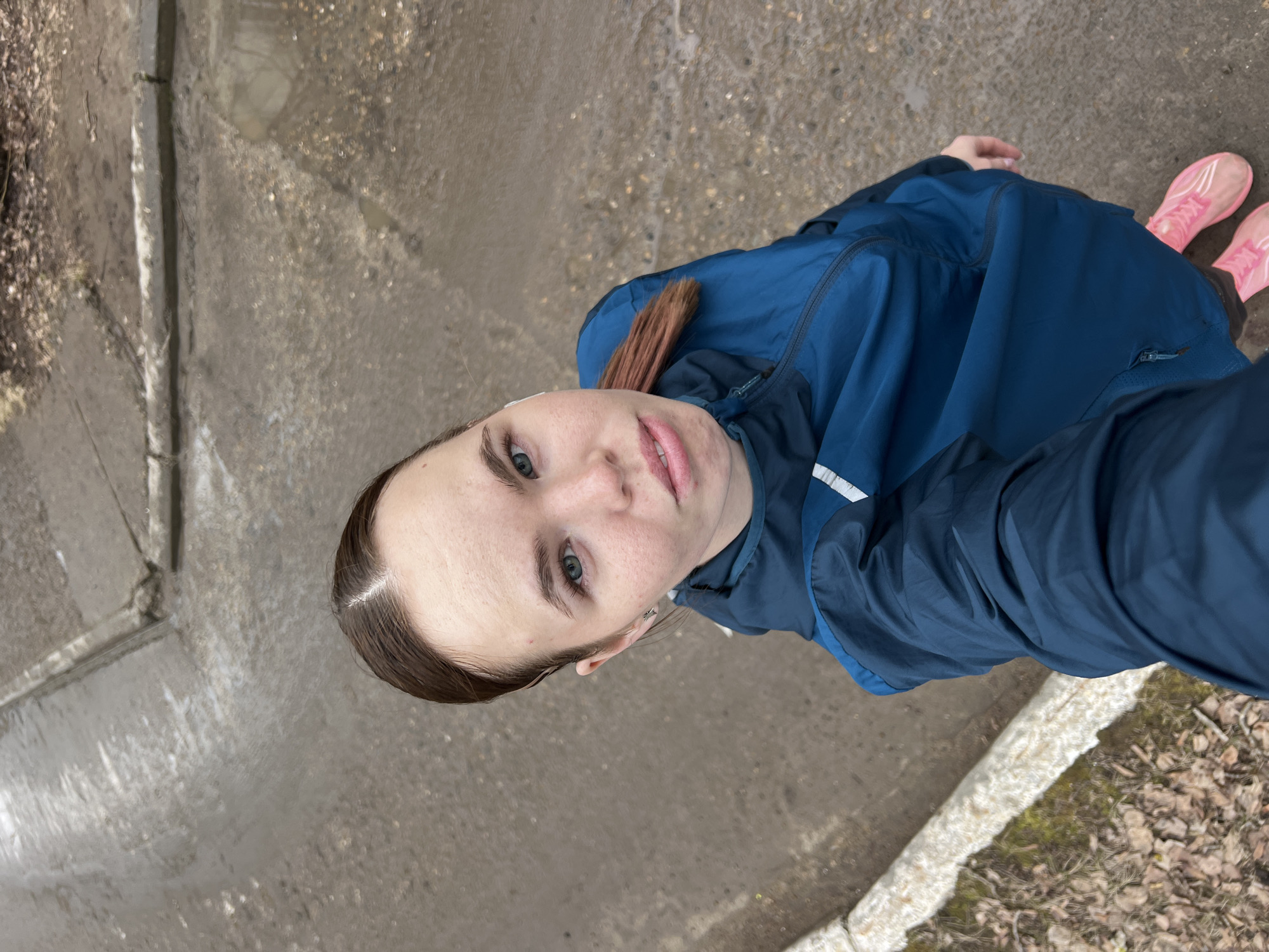 A young Caucasian woman is seen in a selfie taken from a high-angle perspective. She has light skin, blue eyes, and brown hair. She's wearing a blue windbreaker and pink running shoes. The background appears to be a wet, paved surface, possibly a sidewalk or street, alongside a curb. To the right of the frame is a grassy area with fallen leaves. The photograph was taken outdoors in the city of Chaikovsky, Russia.