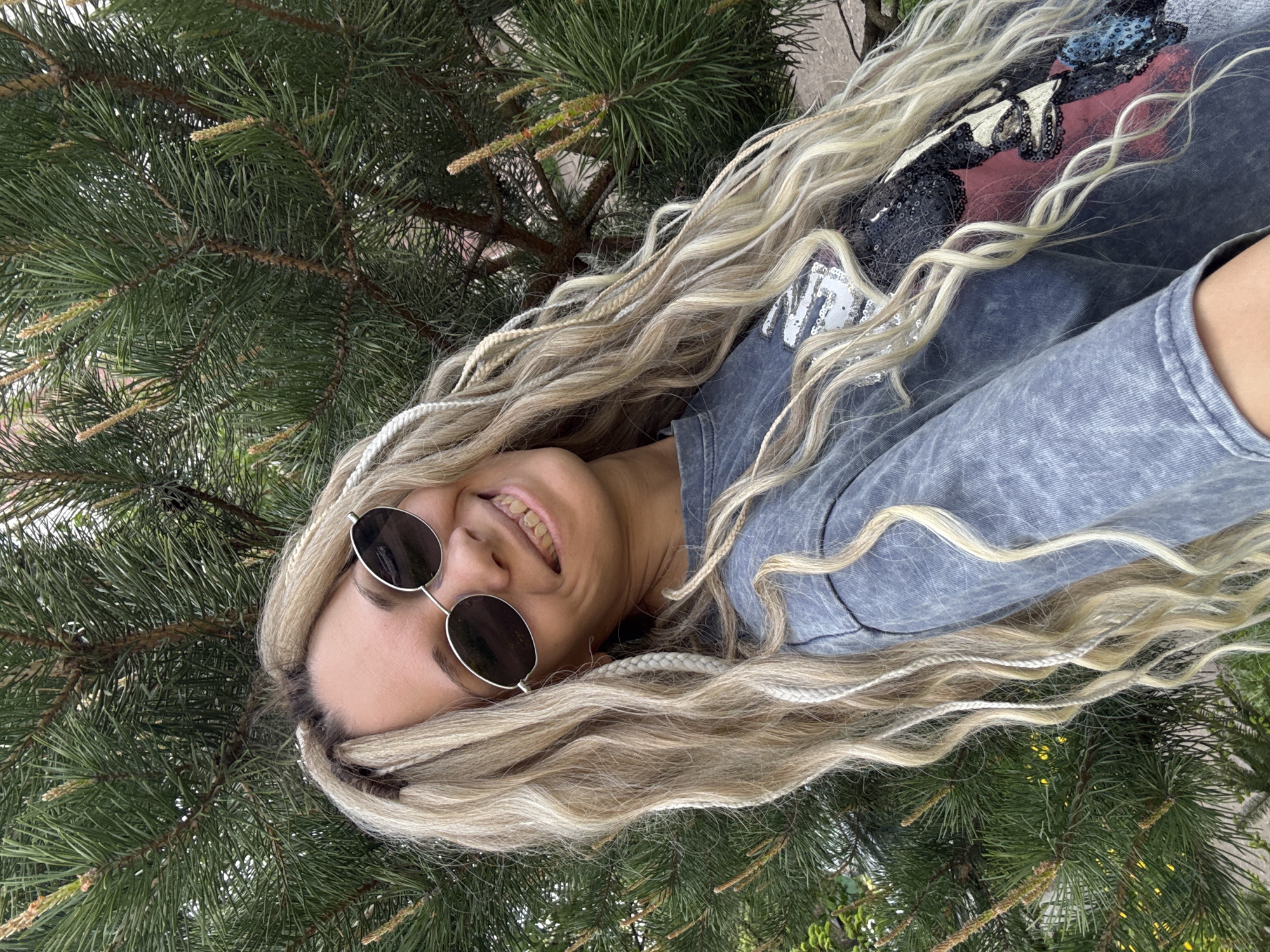 A woman with long blonde braided hair and sunglasses is smiling at the camera. She is wearing a gray shirt with a design on it. She is positioned next to a coniferous tree. This image was taken in Rusanovo, Russia.