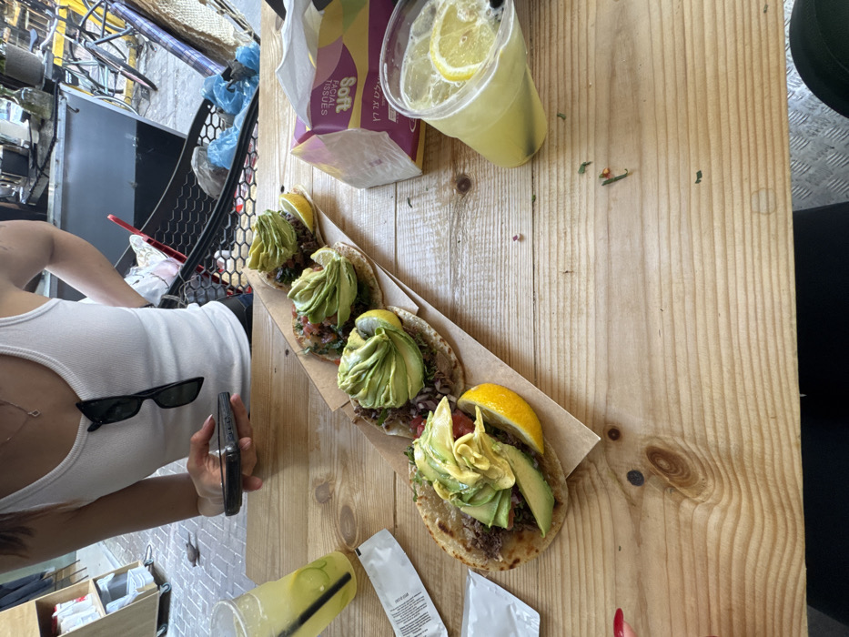 The image captures a casual outdoor dining scene at a wooden plank table in Tel-Aviv, Israel. A person is partially visible in the upper left, wearing a white ribbed tank top with black sunglasses hooked to its collar. This individual holds a black smartphone in their right hand.
Four tacos are arranged in a linear fashion on a strip of brown parchment paper. Each taco consists of a tortilla base topped with dark meat or filling, a red and white diced relish, fanned slices of green avocado, and a yellow lemon wedge garnish.
Two clear plastic cups containing a yellow liquid with lemon slices and ice are present. One cup with a black straw is on the upper left, and another, also with a black straw, is on the right. A white packet rests near the left cup. A box of "Soft Facial Tissues" bearing Hebrew text is positioned on the right side of the table. Small green and red food debris are scattered across the wooden table surface.
The background reveals a paved street or pedestrian area with a black mesh basket containing blue plastic bags and other items, indicative of an urban market or street vendor environment. Parts of a bicycle are visible behind the basket. The ground directly beneath the table in the foreground features a metal diamond plate pattern. The scene depicts a meal in a relaxed, bustling Tel-Aviv urban setting, supported by the visible Hebrew text on the tissue box.