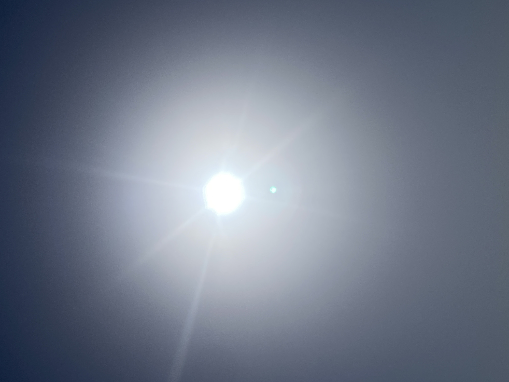 The image shows a bright light source, possibly the sun, dominating the center of the frame. The surrounding sky fades from a dark blue-gray on the edges to a bright white around the light source, obscuring any specific details of the location. There are visible light rays emanating from the central point.