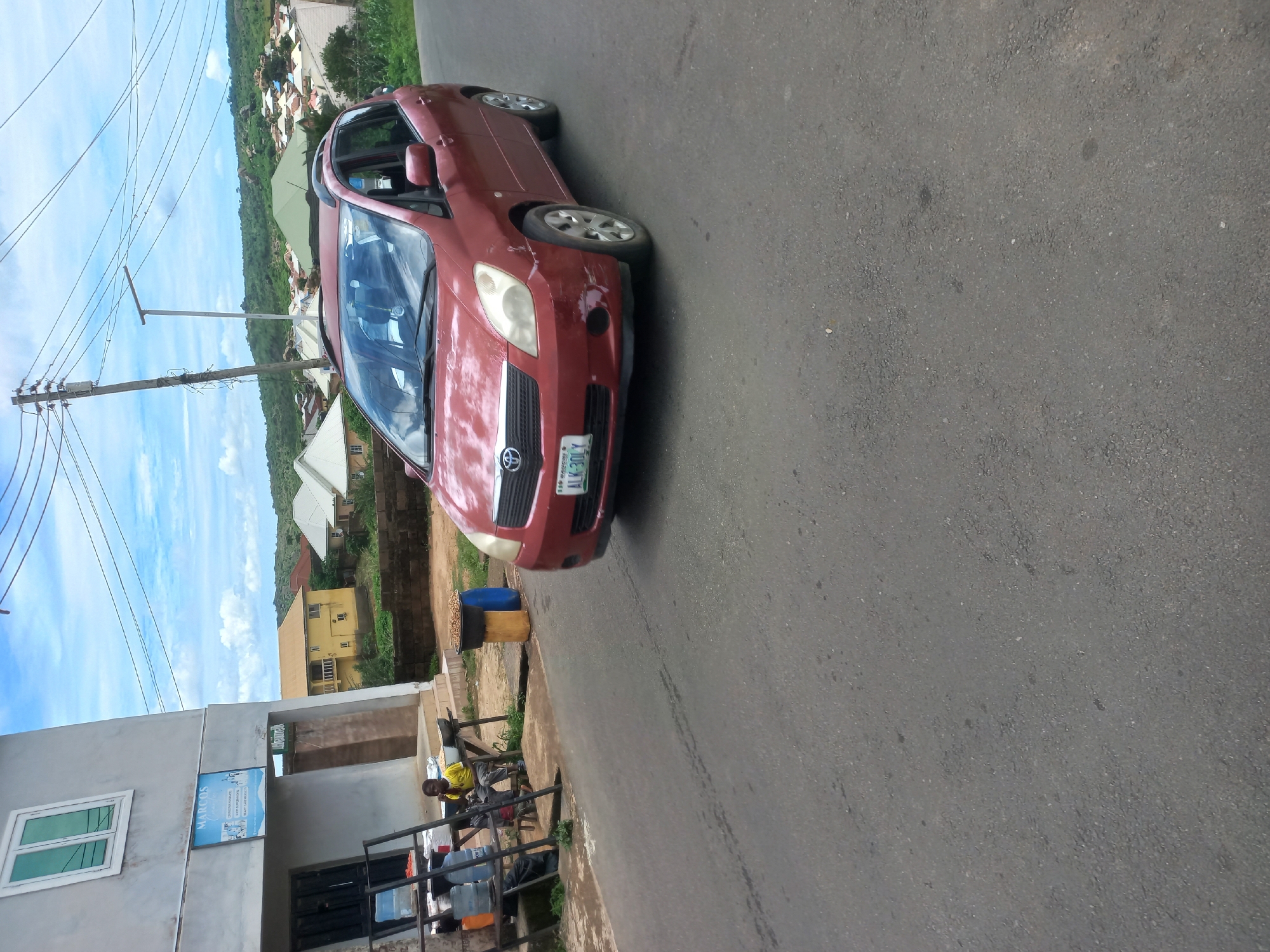 A dark red minivan is parked on a paved road, facing right. The minivan has a license plate that reads "ALK-301LY". To the left of the minivan, a young boy wearing a yellow shirt is seated on a wooden bench in front of a light-colored building. The building has a sign that says "MARCOS". There are several plastic containers and other items on shelves to the left of the boy. In the background, there are houses and trees on a hillside. The sky is blue with some clouds. The scene appears to be in a residential area of Jos, Nigeria, given the architecture and vegetation.