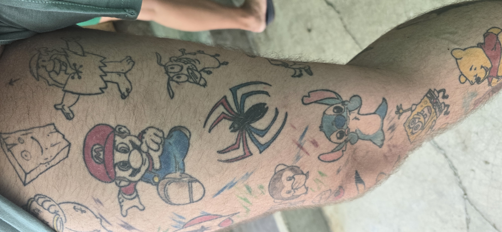 A close-up shot captures a person's leg, likely the upper thigh, heavily adorned with a collection of pop culture-inspired tattoos. The skin is hairy, and the tattoos appear to be in a simple, sketch-like or amateur style, featuring black outlines with some color fill.
Prominently visible characters include:
- **Pebbles Flintstone** in outline style, positioned in the top left.
- **Courage the Cowardly Dog** in outline, lying down, situated above a large spider.
- A colorful **Mario** from Super Mario Bros. with a red cap and blue overalls, located in the center-left.
- A distinct **spider** with a black body and red and blue striped legs, central to the image.
- **Stitch** from Lilo & Stitch, colored light blue and pink, positioned to the right of the spider.
- **SpongeBob SquarePants** in yellow with black outlines, towards the bottom right.
- **Winnie the Pooh** with a yellow body and red shirt, visible in the top right.
- **Plank** from Ed, Edd n Eddy, depicted as a wooden board with a face, in outline style on the bottom left.
- A small, brown and white monkey tattoo is visible below Stitch.
Other smaller, less distinct designs and faint colored scribbles are scattered around these main figures. Green fabric, likely clothing, is visible at the top and bottom left edges of the frame. The background is out of focus, revealing a light-colored, possibly concrete or paved surface with subtle lines, suggesting an outdoor or patio setting during daytime. No discernible text is visible within the tattoos or the background. The scene is located in Govedare, Bulgaria.