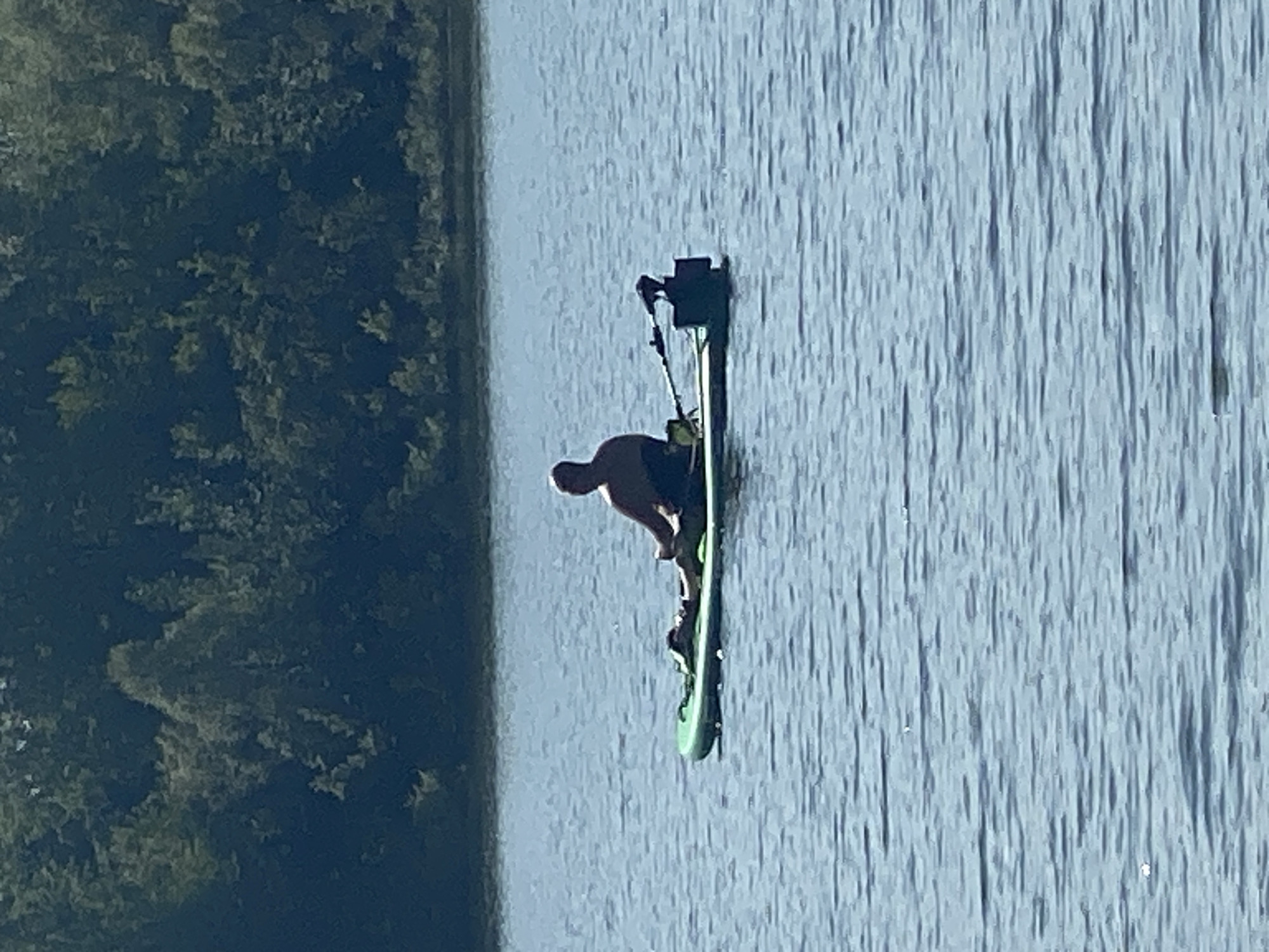 A person is paddleboarding on a body of water. The person is sitting on a green paddleboard and appears to be resting. In the background, there is a densely wooded area. The image is taken from a distance, making it difficult to ascertain specific details about the person's attire or facial features. The water is calm with ripples. The overall scene suggests a tranquil day outdoors.