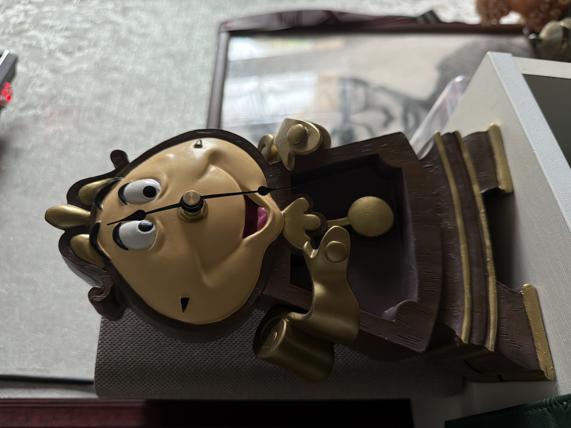 The image shows a decorative clock, designed to resemble the character Lumière from Disney's Beauty and the Beast, placed on a white shelf. The clock is brown with gold accents and has a human-like face with wide eyes and an open mouth. Its pendulum is visible, hanging below its chin. In the background, a framed black and white portrait of a person is partially visible. To the left of the clock, a textured grey object, possibly a speaker or lampshade, is present. The scene is indoors, with a textured wall in the background. There is no indication of the city of Александровка, Russia, in the image.