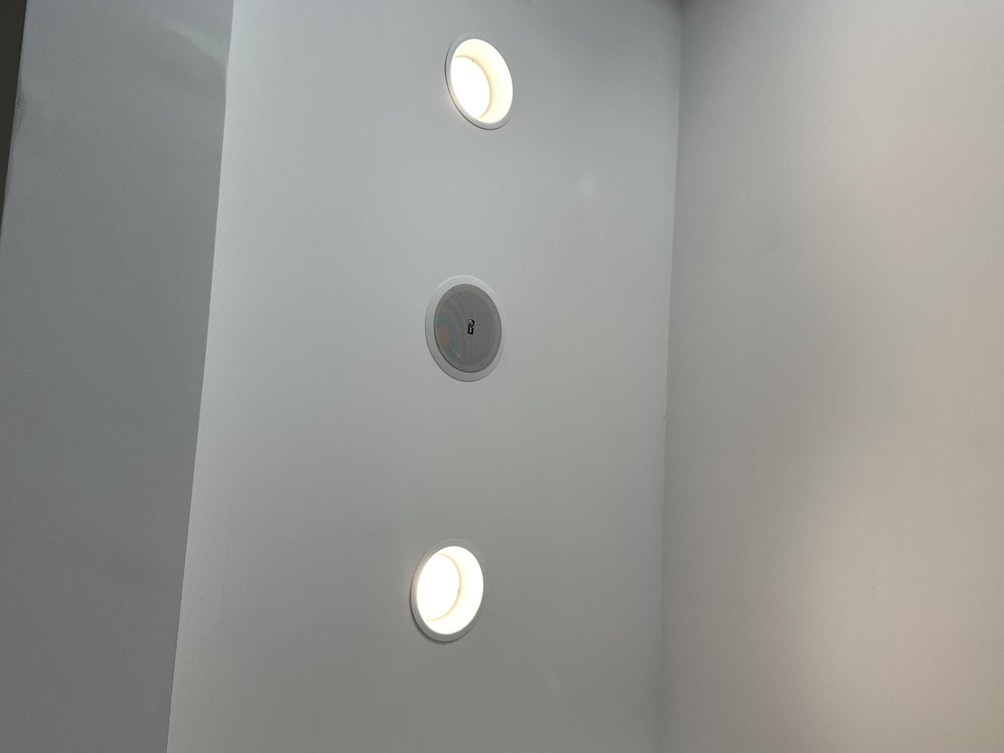 The image shows a ceiling with three recessed fixtures. Two of the fixtures are circular lights emitting a warm yellow glow, and the third is a round speaker with a white grille and a black logo. The ceiling itself is a matte white surface, with a subtle shadow line separating it from a darker upper section, possibly a different ceiling plane or structural element. The lighting suggests an indoor setting, likely a residential or commercial space. The location in Boreti, Montenegro, implies it is part of a building within that coastal town. No people or specific actions are depicted.