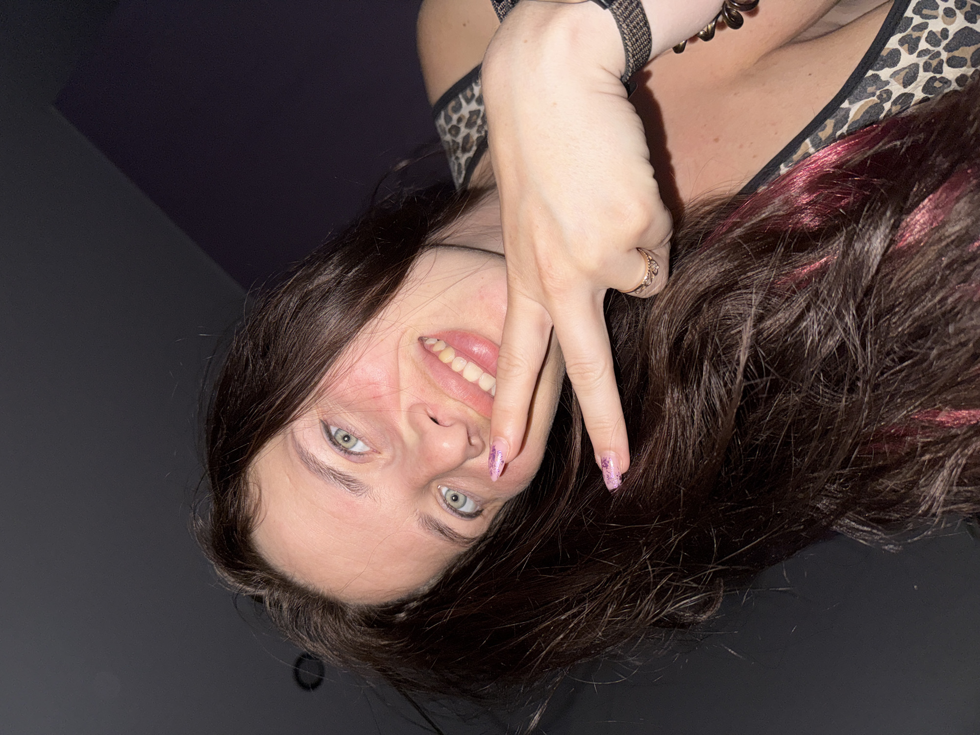 A young woman with long, dark hair and light green eyes is looking at the camera and smiling. She is wearing a leopard-print top. Her right hand is raised to her face, and she is making a peace sign with her fingers. She has pink nail polish on her fingernails, which have glitter on them. She is also wearing a ring on her right hand. The background is dark and out of focus, with a hint of purple in the upper right corner, suggesting an indoor setting.