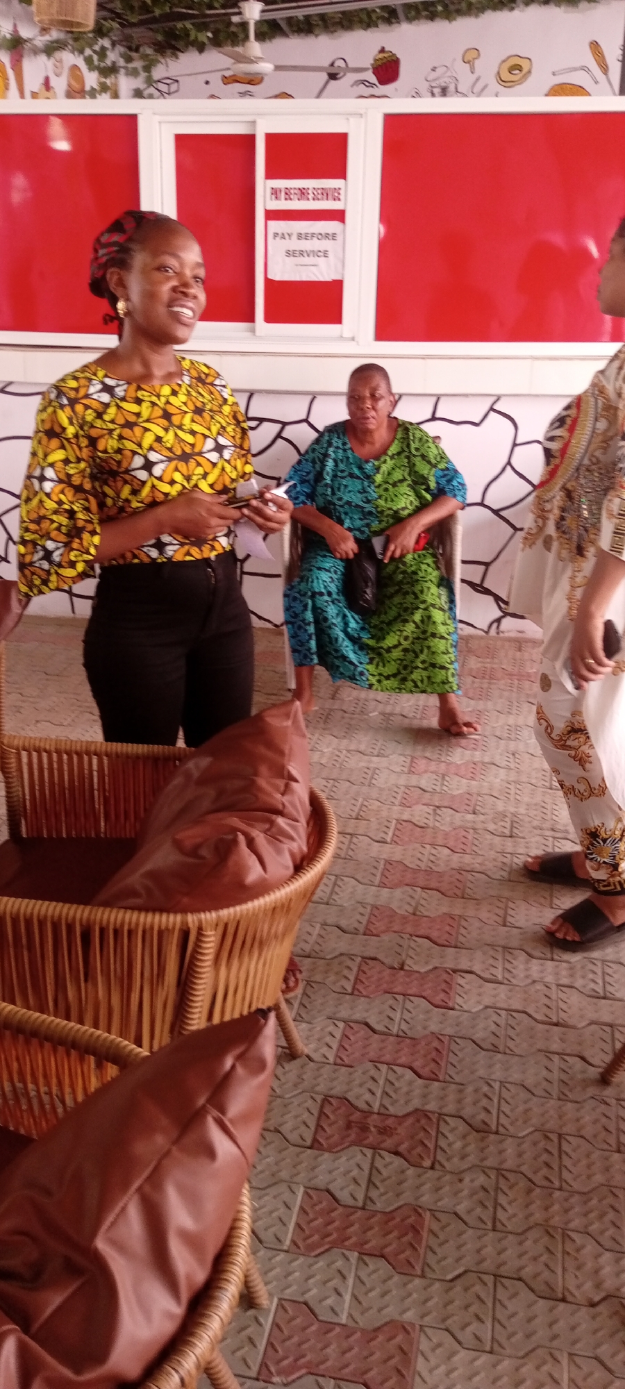 The image shows a partially indoor setting in Sabon Gari East, Nigeria. Three women are visible. One woman, wearing a yellow patterned top and black pants, is standing and holding a mobile phone. Another woman, wearing a green and blue patterned dress, is seated on a chair. A third woman is partially visible on the right, wearing a patterned white outfit. In the background, there is a red wall with a "PAY BEFORE SERVICE" sign. There are wicker chairs with brown cushions in the foreground. The floor is paved with patterned tiles. The background wall also has cartoonish food illustrations. A ceiling fan is visible above.