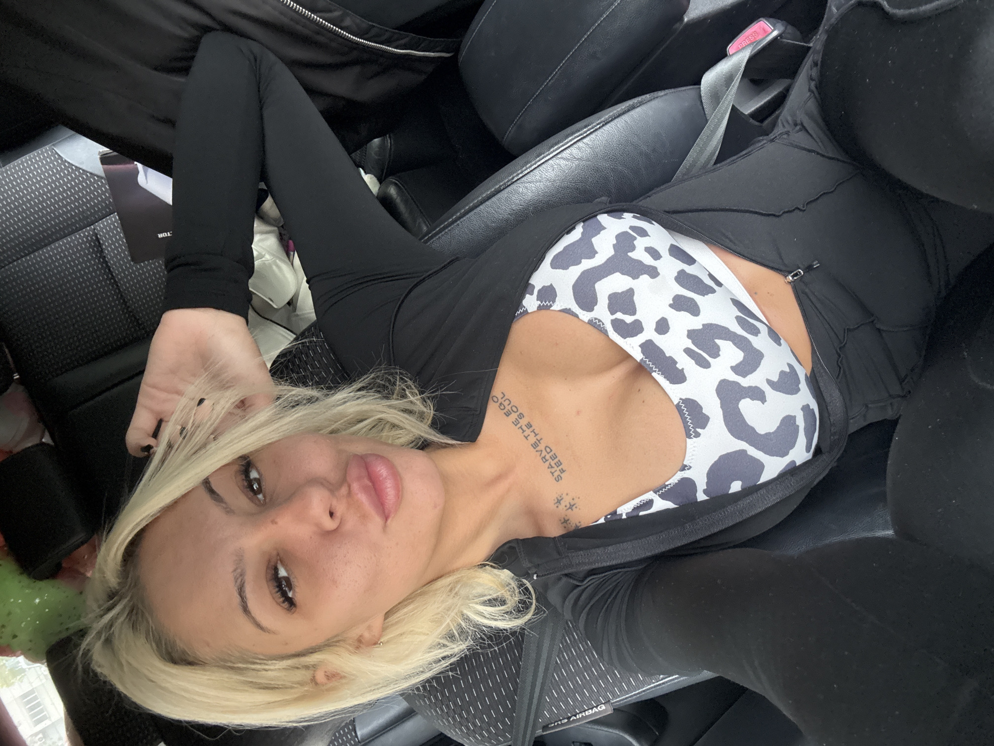 The image features a fair-skinned woman with long blonde hair, lying on her back inside what appears to be a car. She looks directly at the camera with a neutral expression, her lips slightly parted. She is wearing a black long-sleeved athletic top, unzipped to reveal a white and dark gray animal-print sports bra. Her bottom half is clad in black leggings.
A notable detail is a tattoo on her chest/collarbone area which reads "STARVE THE EGO FEED THE SOUL" in capital letters, with a series of small star designs underneath. The setting is clearly a car interior, indicated by the black leather seats, a gray seatbelt with a visible "PRESS" buckle, and the "SRS AIRBAG" label on the seat cushion. Part of a dark object or magazine with the text "TOR" is visible near her left leg. The scene is brightly lit, suggesting it is daytime. The image captures a casual self-portrait moment, taken in Plovdiv, Bulgaria.