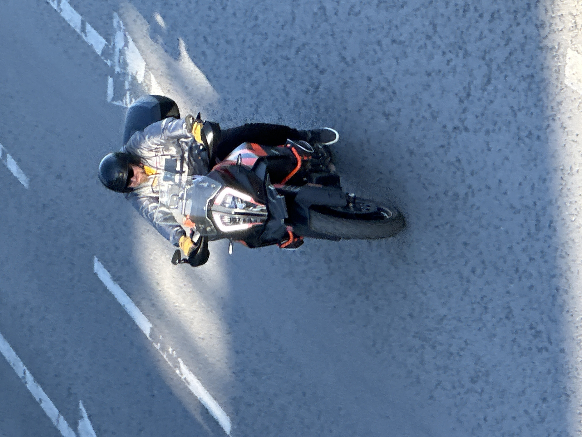 An aerial perspective captures a motorcyclist traveling on a road marked with white lines, likely in Veliko Tarnovo, Bulgaria, given the provided context. The rider, wearing a helmet and silver jacket, is positioned on a dark-colored motorcycle with orange accents. The road surface is a speckled gray. The light direction creates highlights and shadows on the road.