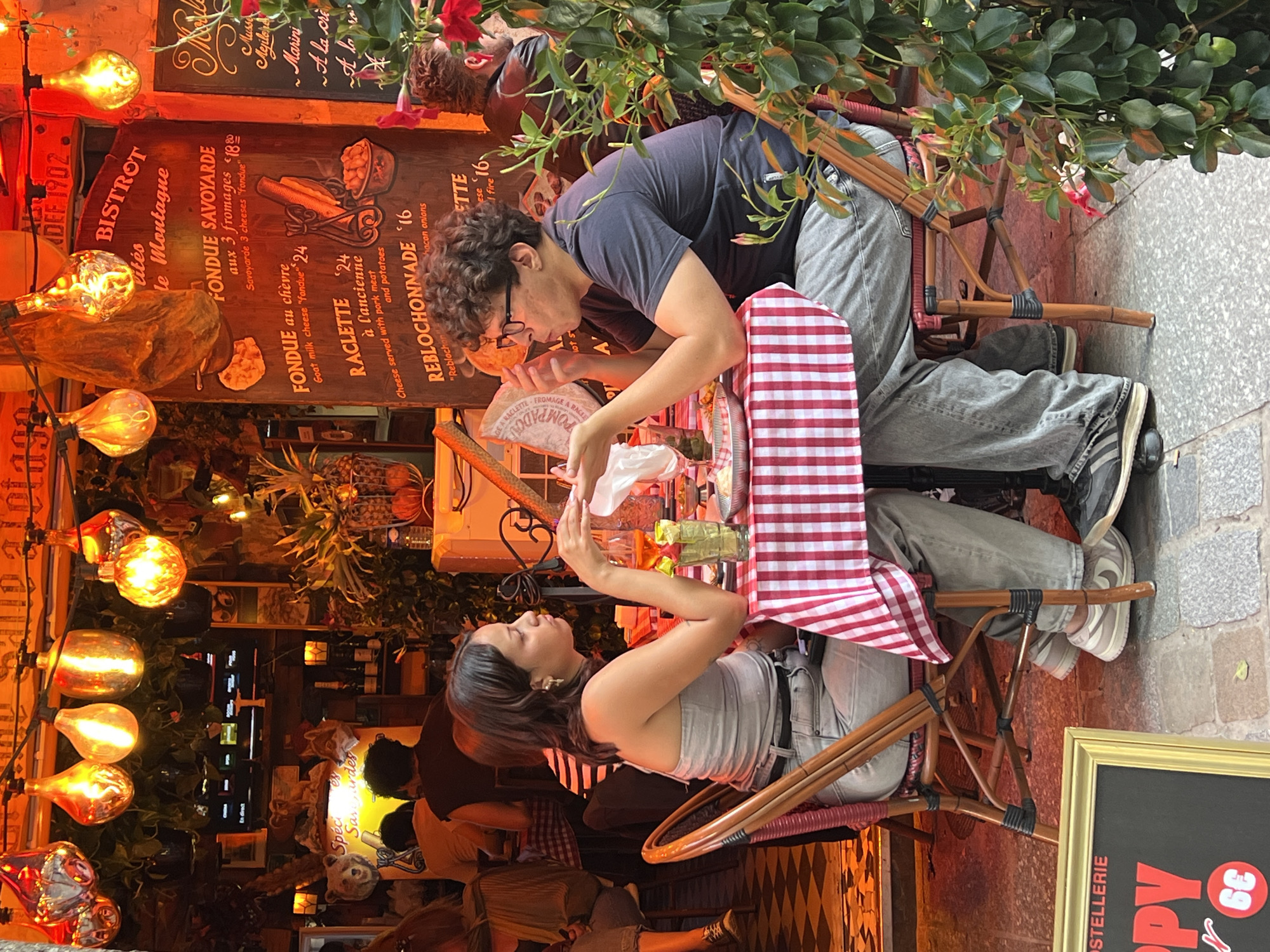A young couple is seated at a table outside a Parisian bistro, possibly Le Montaigne, indicated by the signage. The woman, wearing a grey strapless top and light-colored jeans, sits on the left, her left arm resting on the table. The man, in a dark t-shirt and grey pants, sits opposite her, gesturing with his hands. Both are looking at each other and appear engaged in conversation. The table is covered with a red and white checkered tablecloth, laden with plates, glasses, and what appears to be a carafe of orange liquid. The bistro's exterior is decorated with warm, ambient lighting from large Edison-style bulbs hanging overhead and foliage adorns the scene. The man is wearing glasses and has curly hair. The woman has dark hair and is wearing earrings. The background shows more patrons and restaurant decor. The ground is paved with cobblestones and tiles.