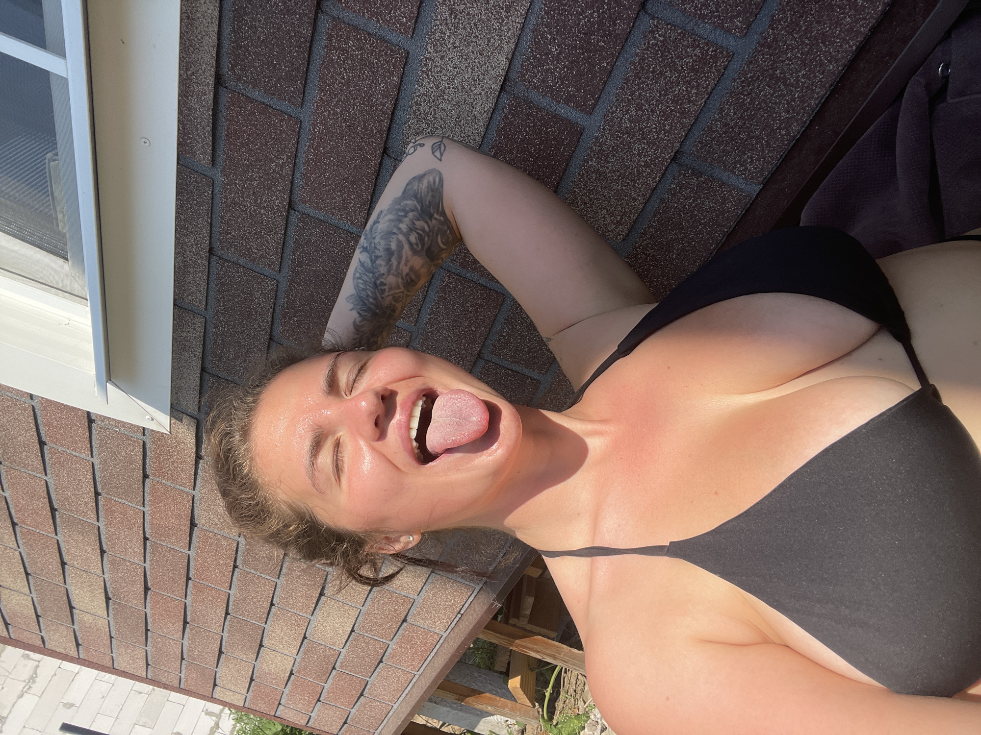 The media file shows a person lying on a roof. The person is wearing a black bikini top and has a tattoo on their arm. Their eyes are closed and their tongue is sticking out. The sun is shining, casting shadows on the roof. The background consists of asphalt shingles and a white window frame. The location context is Мосметаллоконструкция Онт, Russia. The image appears to be taken during the day, with bright sunlight suggesting warm weather. The person's expression suggests a playful or relaxed mood. There is no visible text in the image.