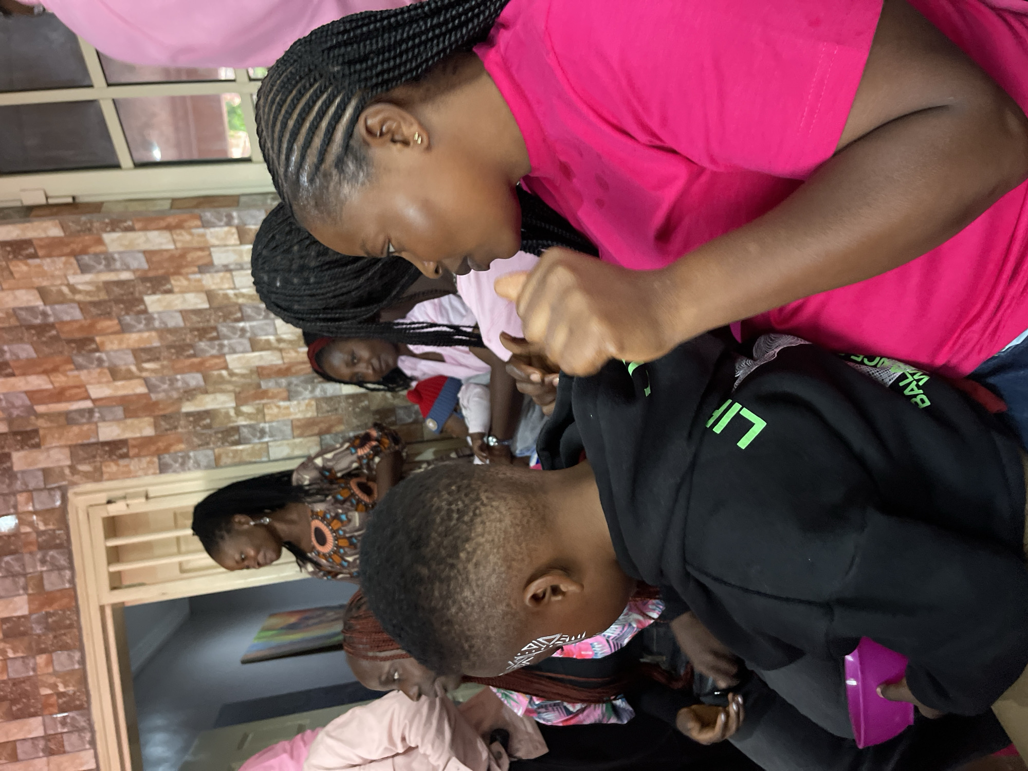The image depicts an indoor gathering of people in what appears to be a home or community space in Gura Top, Nigeria. The foreground features a young Black boy wearing a black hoodie with green lettering, holding a pink bowl in his left hand. He has white face paint on the left side of his face. To his right, a Black woman is visible from the chest up, wearing a bright pink shirt. Her braided hair is pulled back, and she is looking to her left. Behind them, several other individuals are present, including women in patterned clothing and a woman with braided hair wearing a pink shirt. The background shows a textured, tiled wall with earth tones and a doorway with a barred gate. The overall lighting suggests an indoor setting with artificial light contributing to the scene's illumination.
