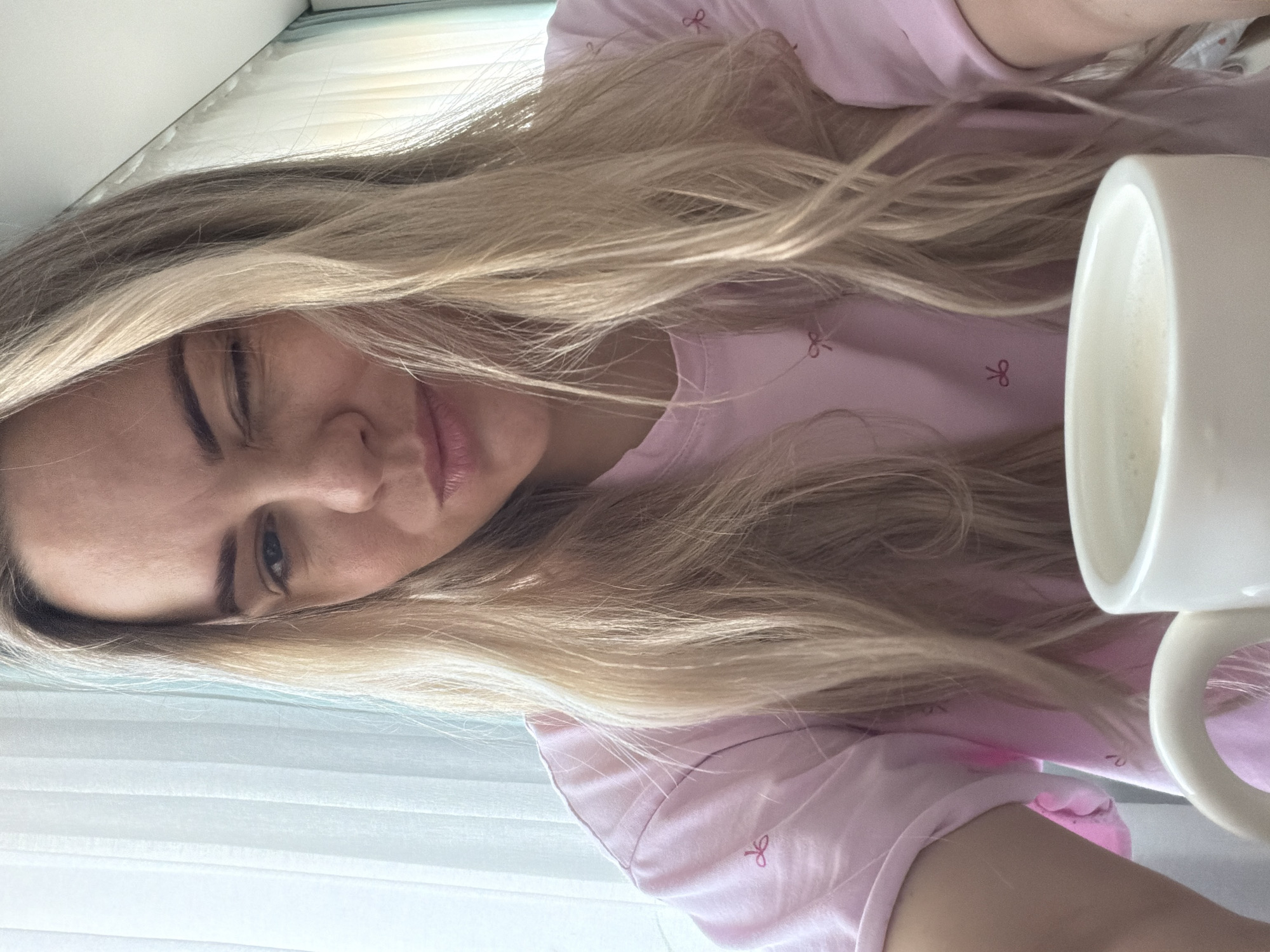 A woman with long blonde hair is indoors, seemingly holding a white mug containing a light-colored liquid, likely a beverage. She is wearing a pink top with a small bow pattern. She winks at the camera. White window blinds are visible in the background. The location is unknown and cannot be identified from the image.