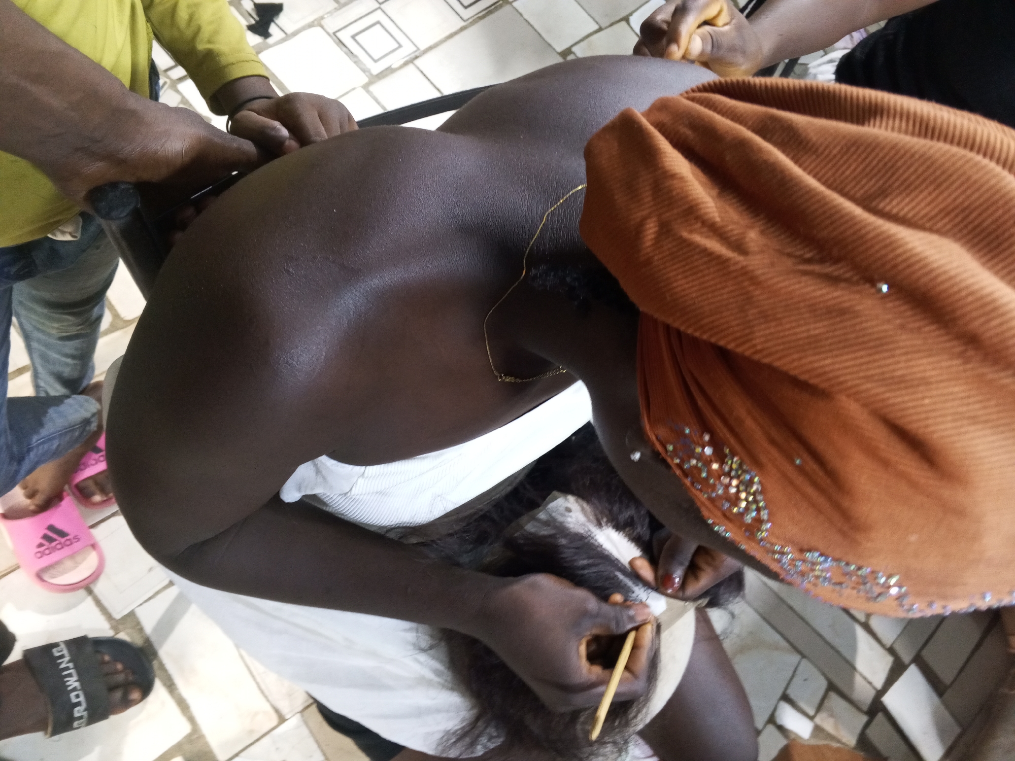 A woman wearing an orange headwrap adorned with shiny beads is shown from an overhead perspective, as she works on a black wig with a wooden tool. Her skin is dark, and she wears a gold chain around her neck. She is seated in front of a white cloth. Several people are standing around her, some holding the wig or her head. One person is wearing a yellow long-sleeved shirt and jeans, and another has on pink Adidas sandals. The floor is tiled in a geometric pattern. The location is Gwong, Nigeria.