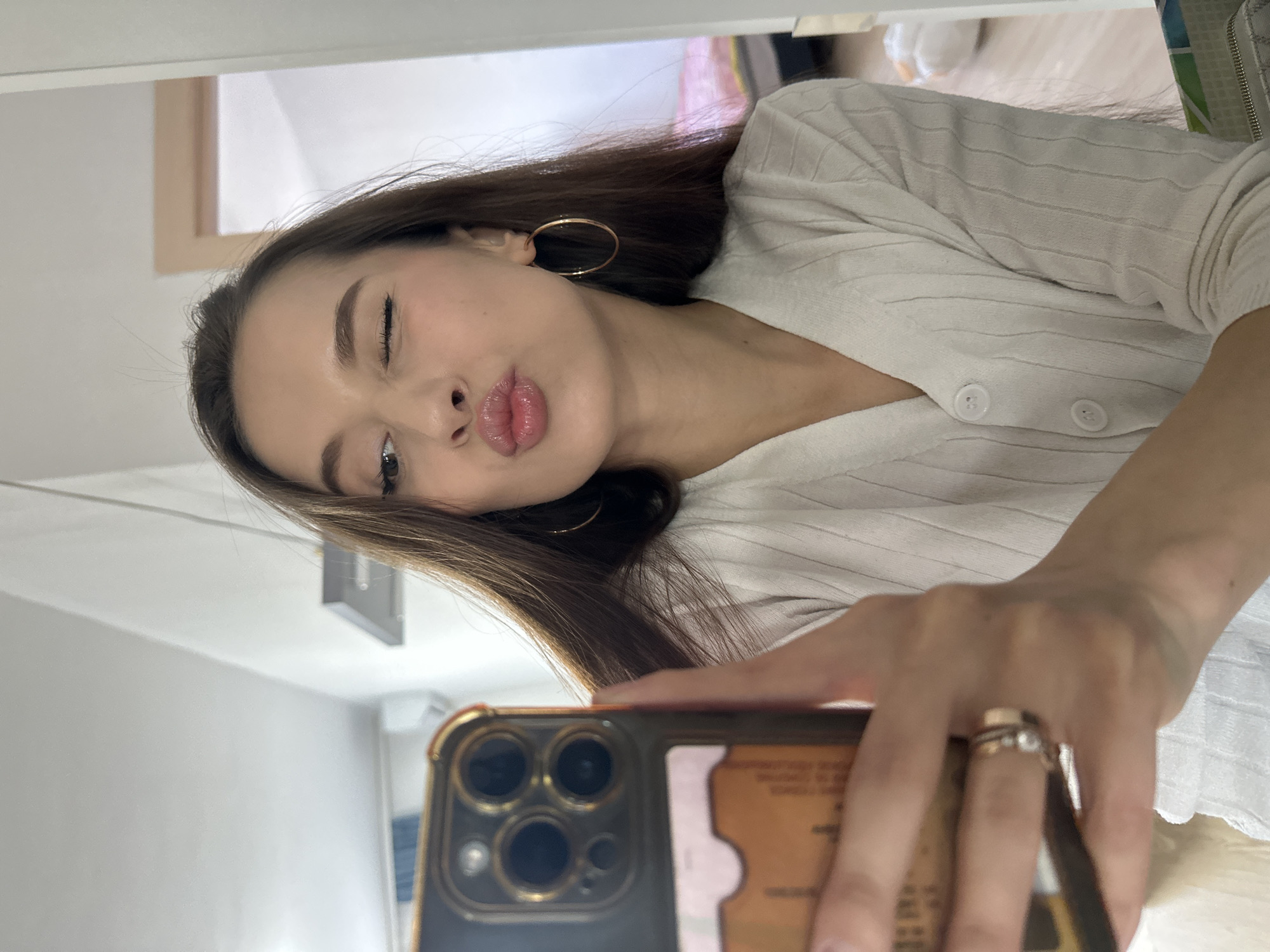 A young woman is captured in a mirror selfie, tilting her head and making a playful expression with one eye winking and her lips pursed in a kiss. She has long, dark brown hair parted on the side and wears a light-colored, possibly cream or white, ribbed knit cardigan with two visible white buttons. Large gold hoop earrings adorn her ears.
Her right hand holds a dark-colored smartphone with a prominent camera array featuring multiple lenses. The phone has a gold-colored frame, and behind it, a light brown card, possibly an ID or bank card, is visible within the phone case. On her ring finger, she wears multiple rings, including a silver-toned band and a gold-toned band with a small clear stone.
The background, seen through the mirror's reflection, appears to be an indoor setting with light-colored walls and ceiling. A light fixture is visible on the ceiling, and a wooden door frame or architectural detail can be seen in the upper left. The overall impression is a casual, indoor moment, likely in a private residence in Yekaterinburg, Russia. The lighting is bright and even, suggesting either a well-lit room during the day or an interior with good artificial lighting. The woman's expression conveys a cheerful and confident mood. Some indistinct text is visible on the card in the phone case.