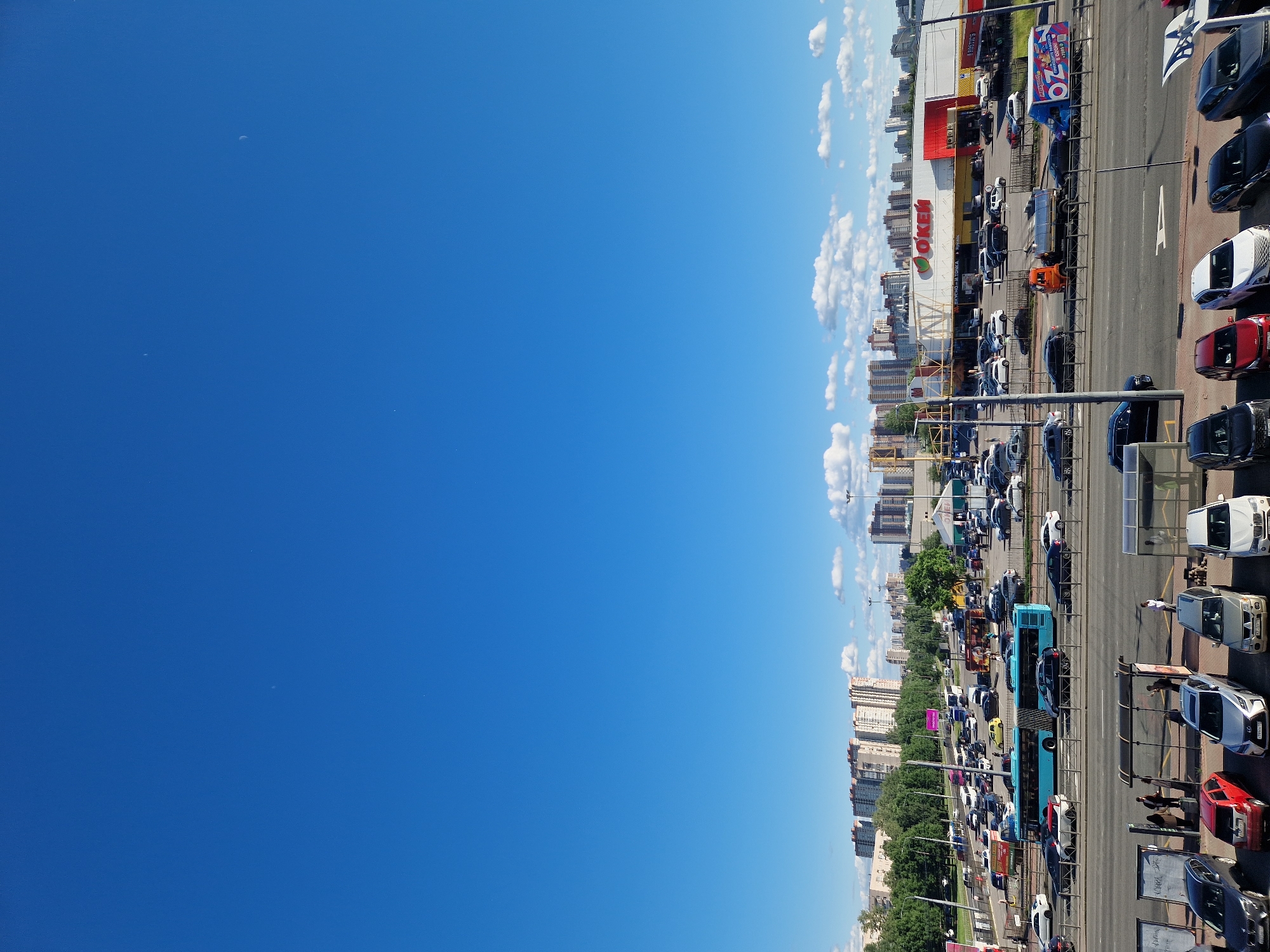 The image captures a wide shot of a city scene on a clear day. The sky is a vibrant blue with a few scattered white clouds. In the foreground, a busy road with parked cars and a bus stop is visible. Several parked cars of various colors and models are lined up. People can be seen at the bus stop, with some standing under the shelters and others walking. Beyond the road, a large parking lot is filled with numerous cars. A blue bus is driving on the road, and another is parked. In the background, a large supermarket with a red and white facade is prominent, along with several other buildings and apartment complexes, indicating an urban setting. Trees and greenery are interspersed throughout the cityscape. The overall impression is a snapshot of everyday urban life with traffic, commerce, and residential areas.