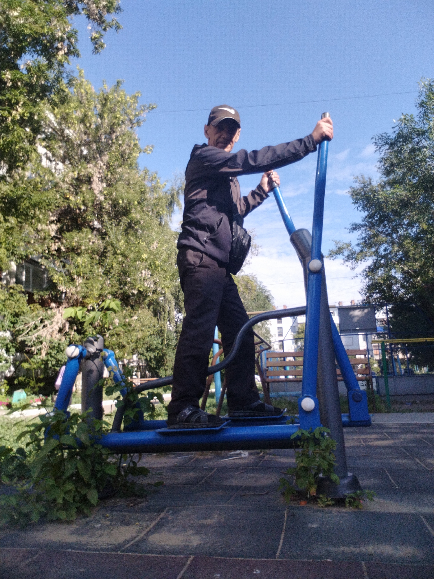 A man wearing a dark jacket, dark pants, and a baseball cap is exercising on a blue outdoor elliptical machine. He is holding the handles and appears to be in motion. The machine is located on a rubberized playground surface. In the background, there are trees, buildings, and a bright blue sky. The scene appears to be a public park or playground in the city of Kostanay, Kazakhstan.